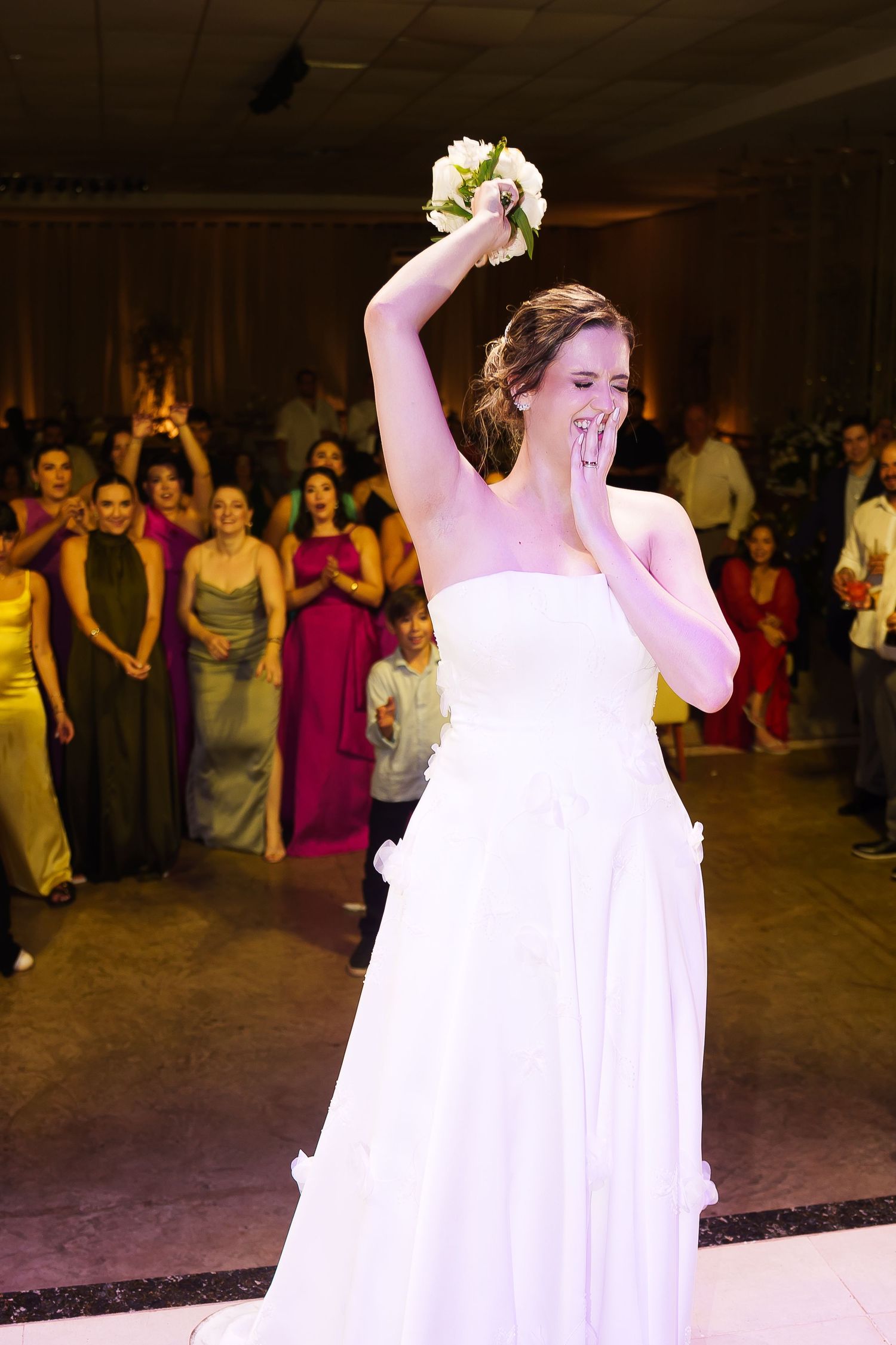 Bride in white strapless gown raising bouquet overhead before toss at wedding reception.