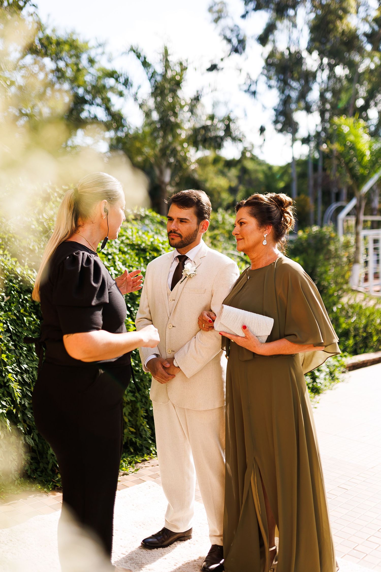 Groom in ivory suit converses with two women in formal attire at an outdoor wedding venue surrounded by lush greenery.