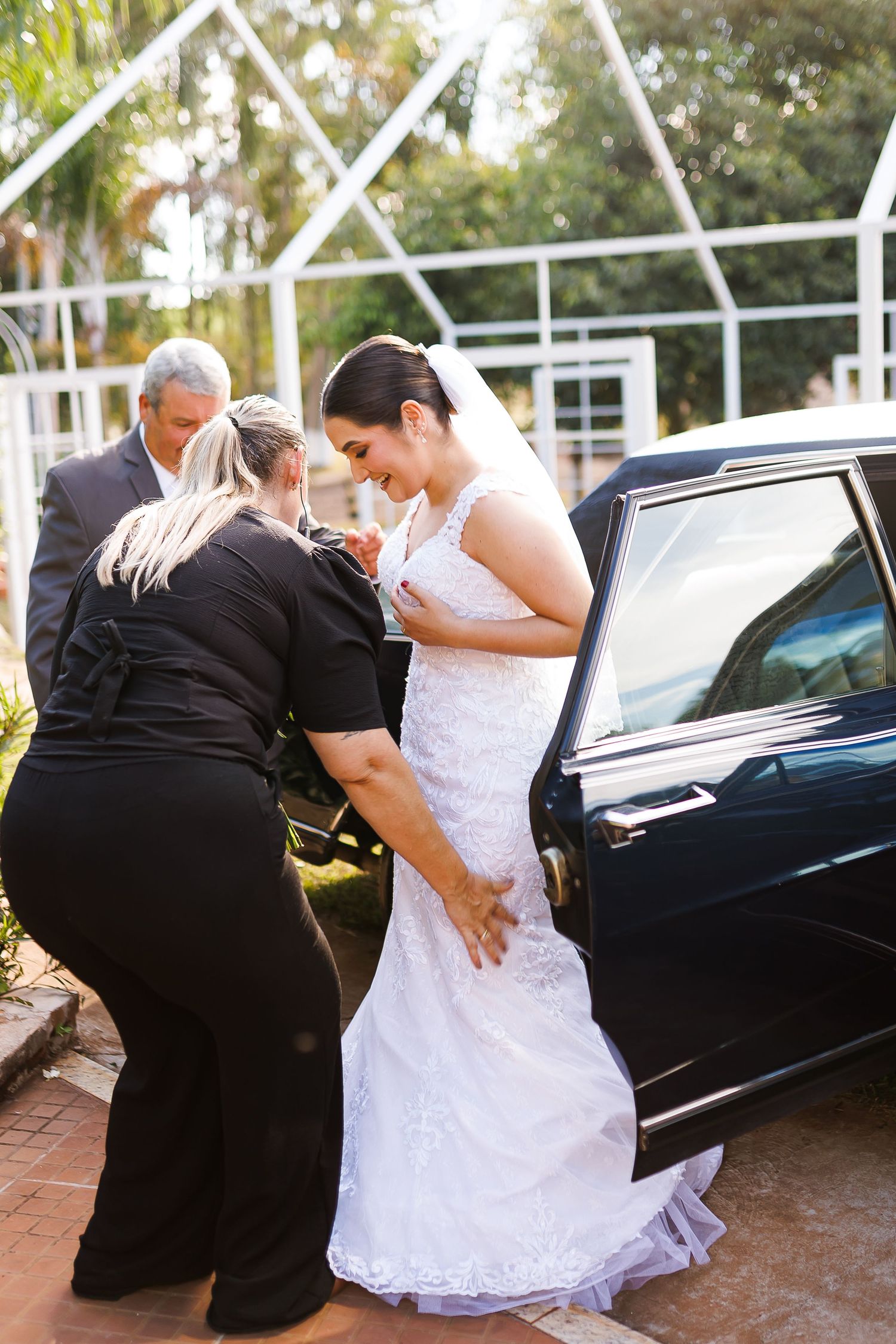 Bride in white lace gown being helped by attendants beside a black car outdoors before her wedding ceremony.