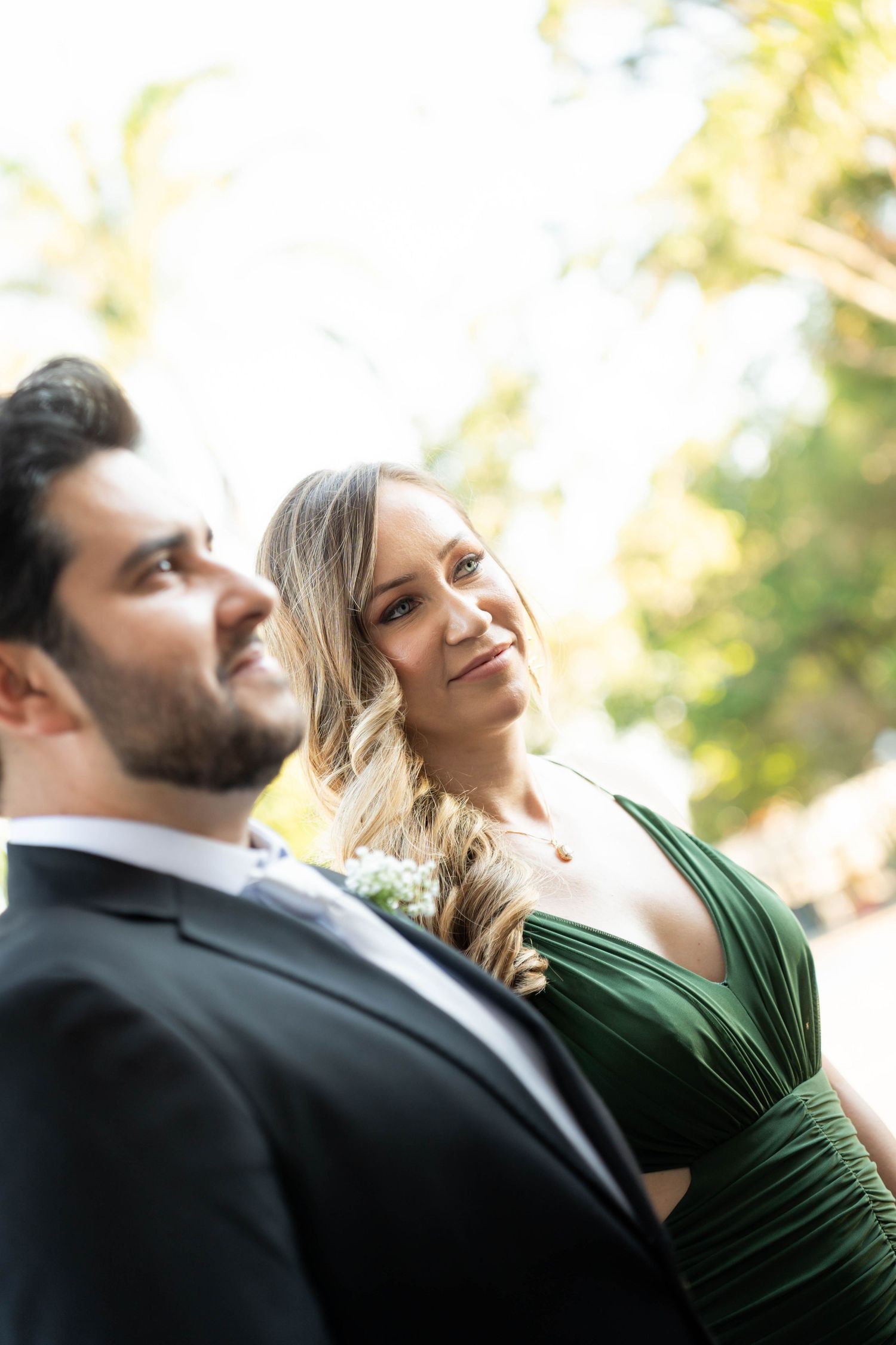 Couple in formal attire, woman in green dress smiling at man in black suit at outdoor wedding.