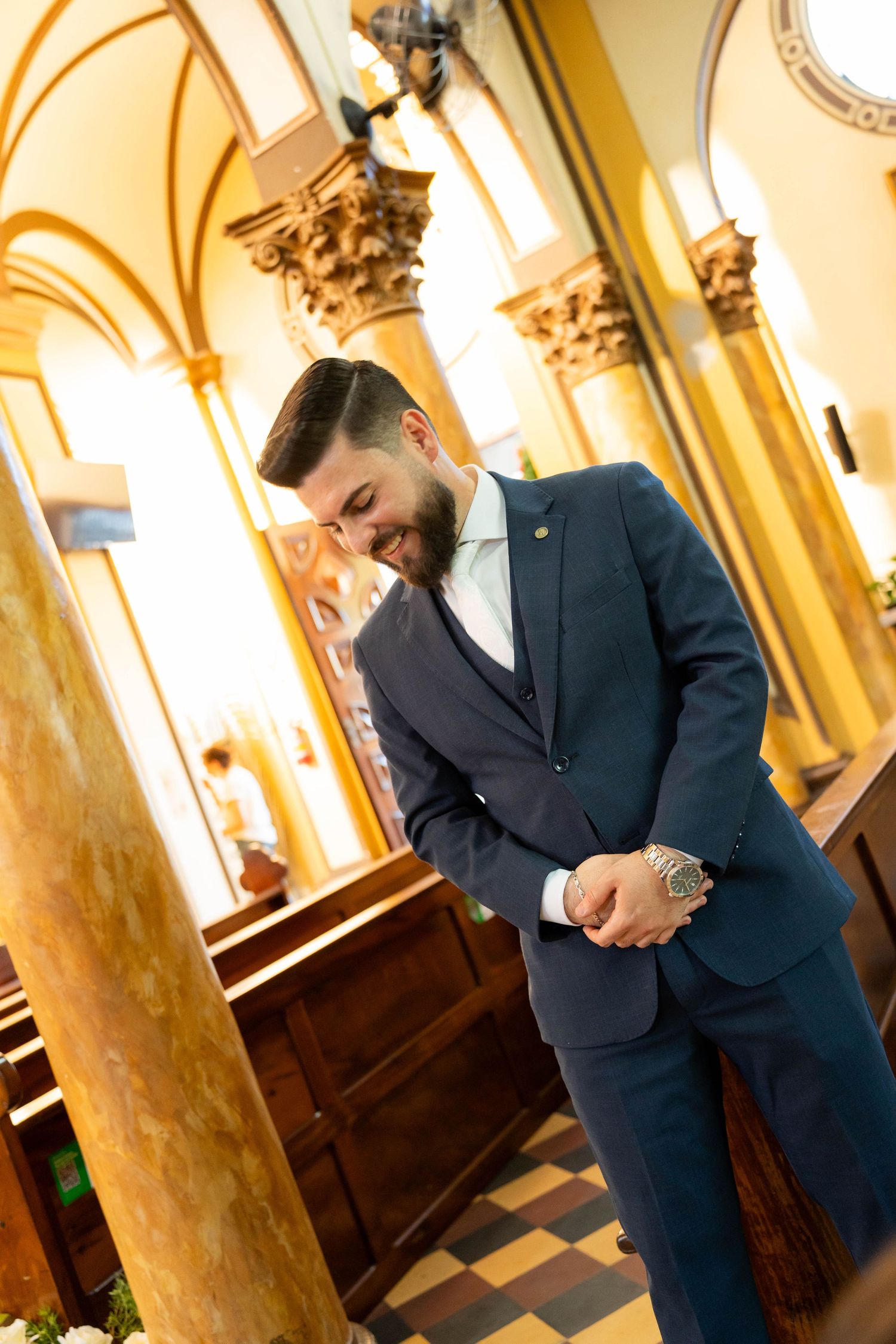 A smiling groom in a navy blue suit stands in an elegant church with ornate columns before his wedding ceremony.