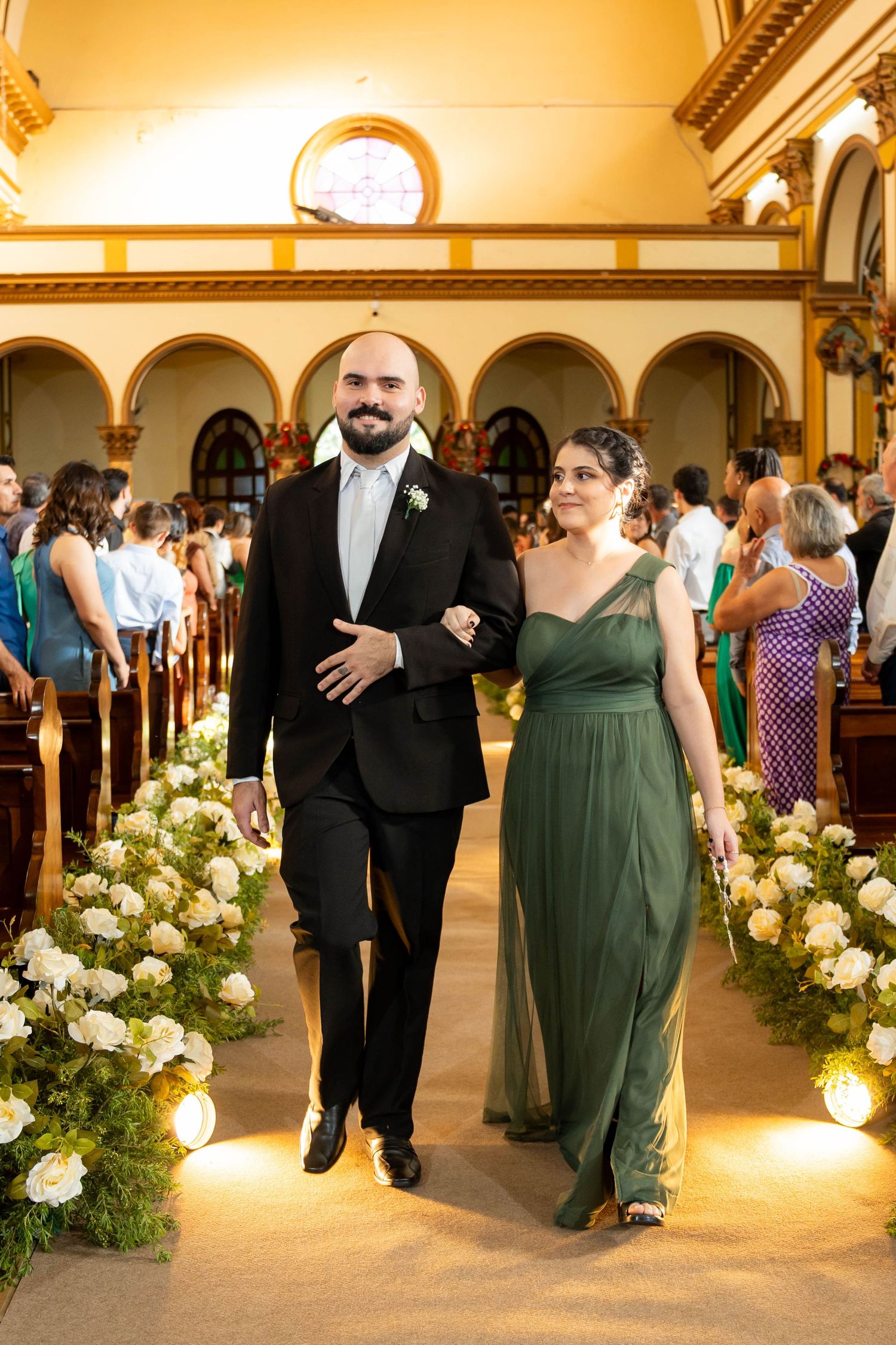 A groom in a black suit and bridesmaid in a green gown walk down a floral-lined church aisle at a wedding.