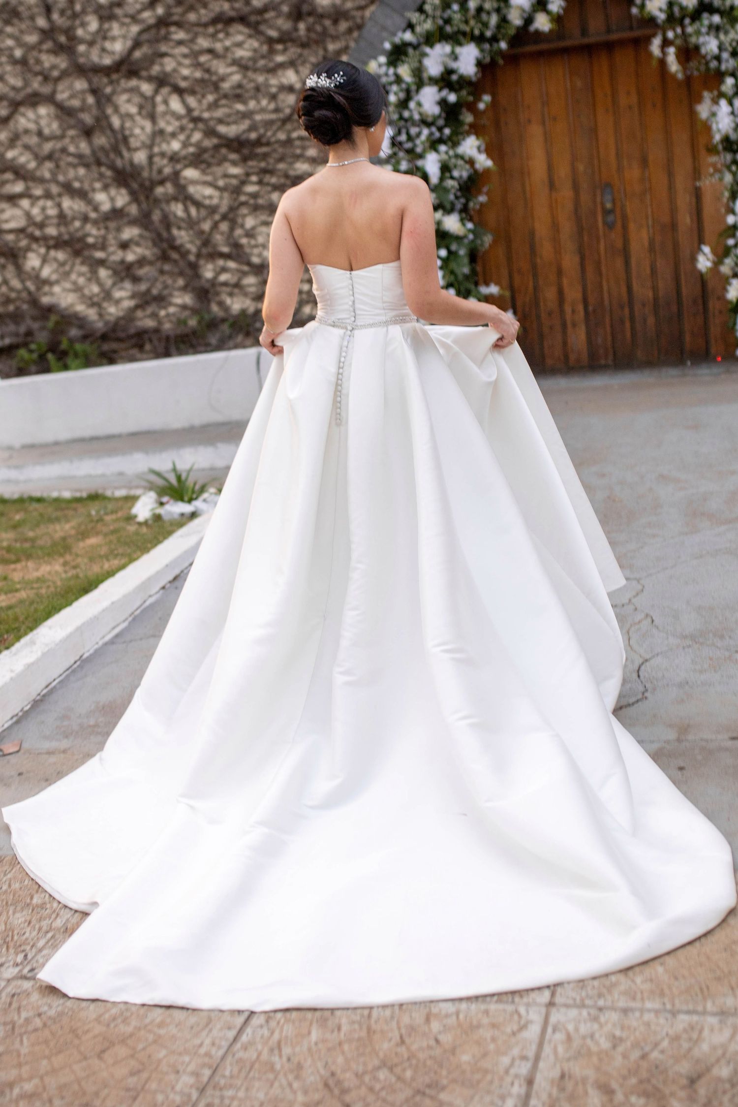 Bride in a strapless white ballgown wedding dress with a long train, viewed from behind, holding her skirt.