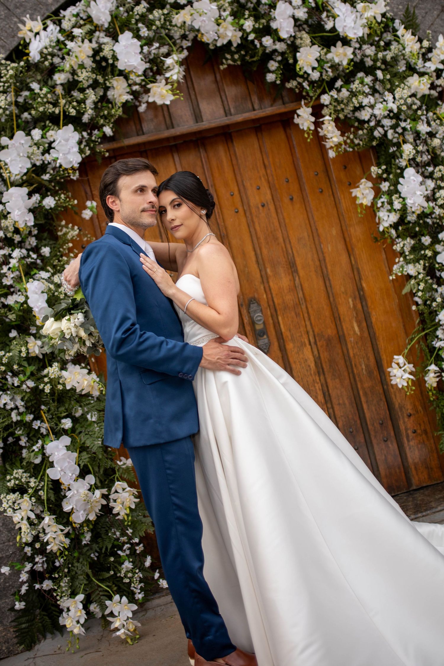 Bride and groom embrace before a wooden door framed by a white floral arch on their wedding day.