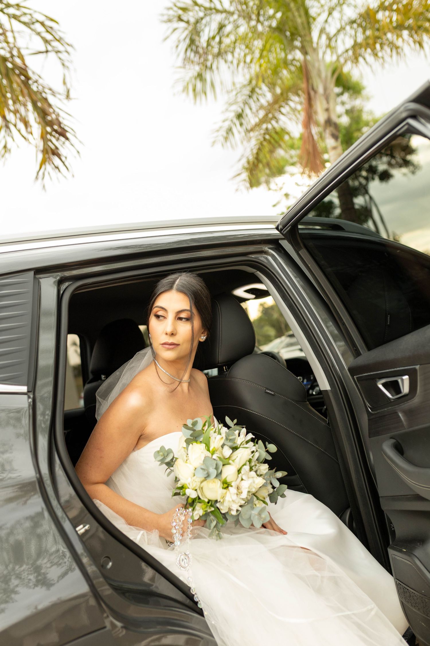 Bride in white strapless gown holding bouquet of white roses stepping out of black luxury car on wedding day.