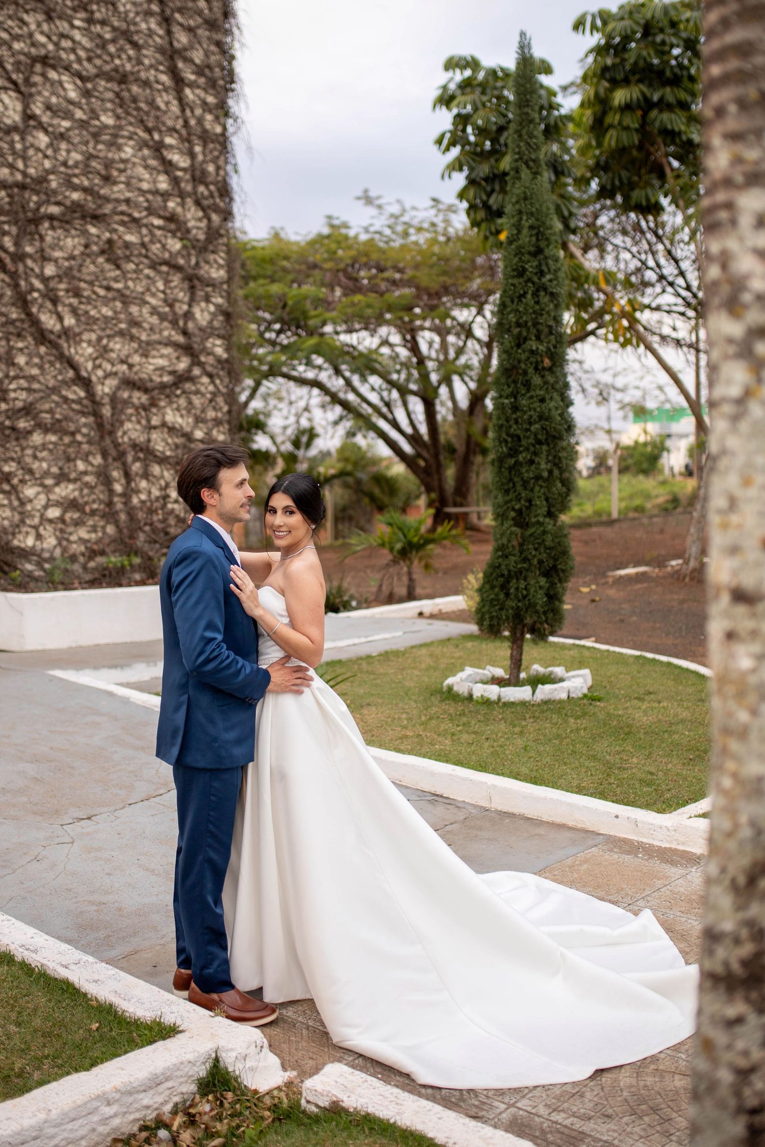 Bride and groom embrace outdoors in a lush garden, she wears a white gown with a long train, he wears a blue suit.