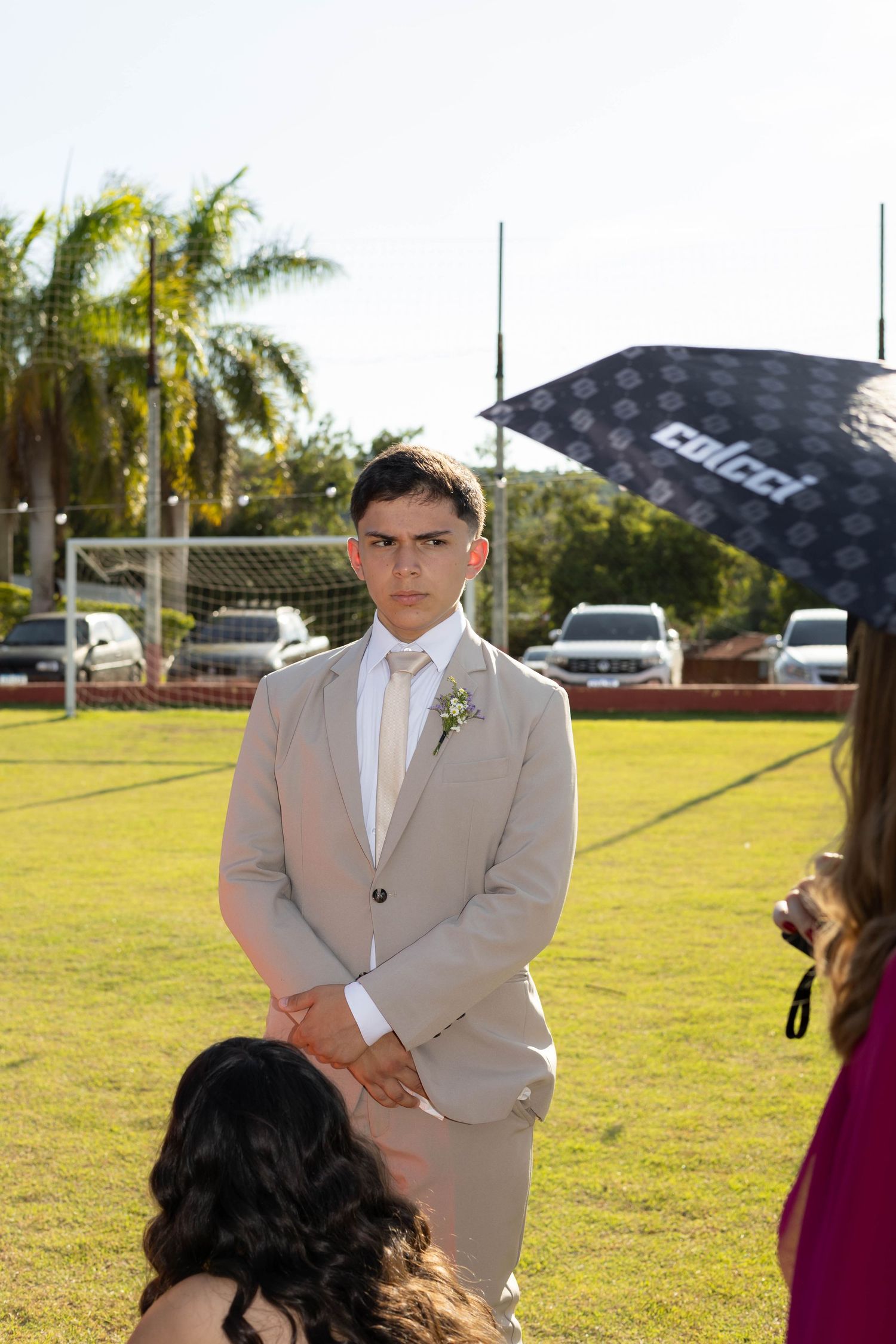 Young man in beige suit with boutonniere stands on green lawn at outdoor formal event with palm trees.