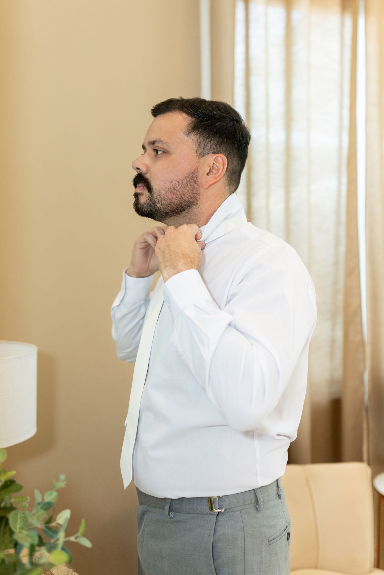 Bearded man in white dress shirt and tie adjusting his collar while getting dressed in a hotel room.