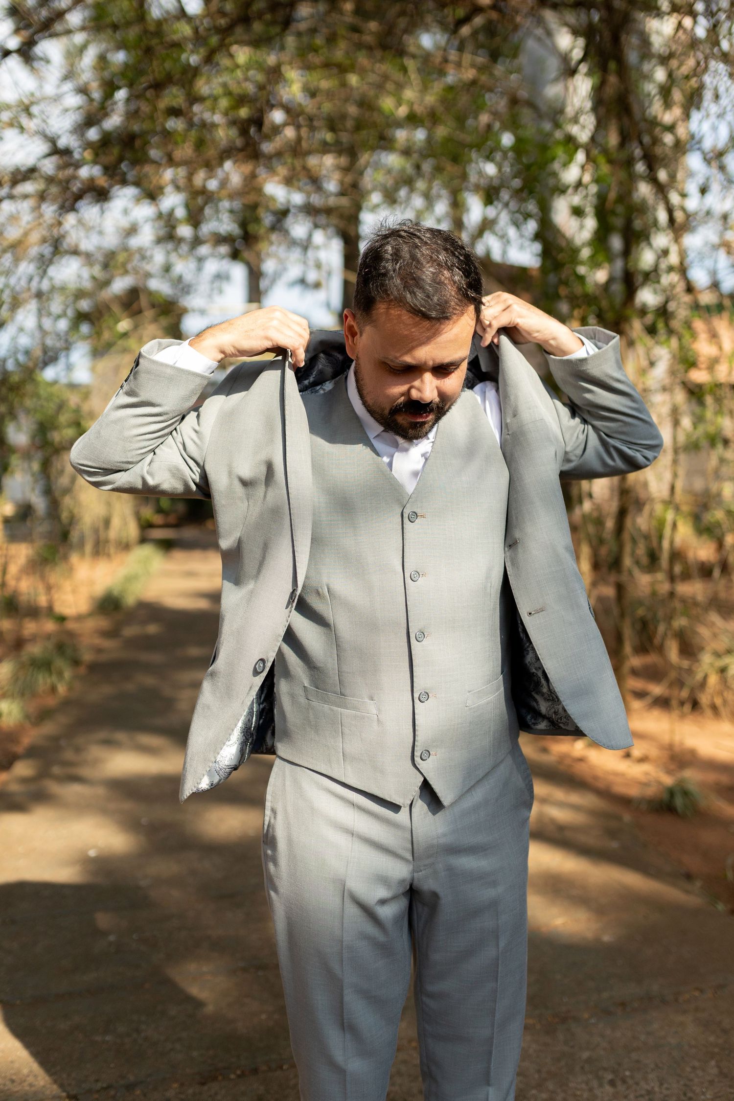 Man in gray three-piece suit adjusting collar outdoors among trees.