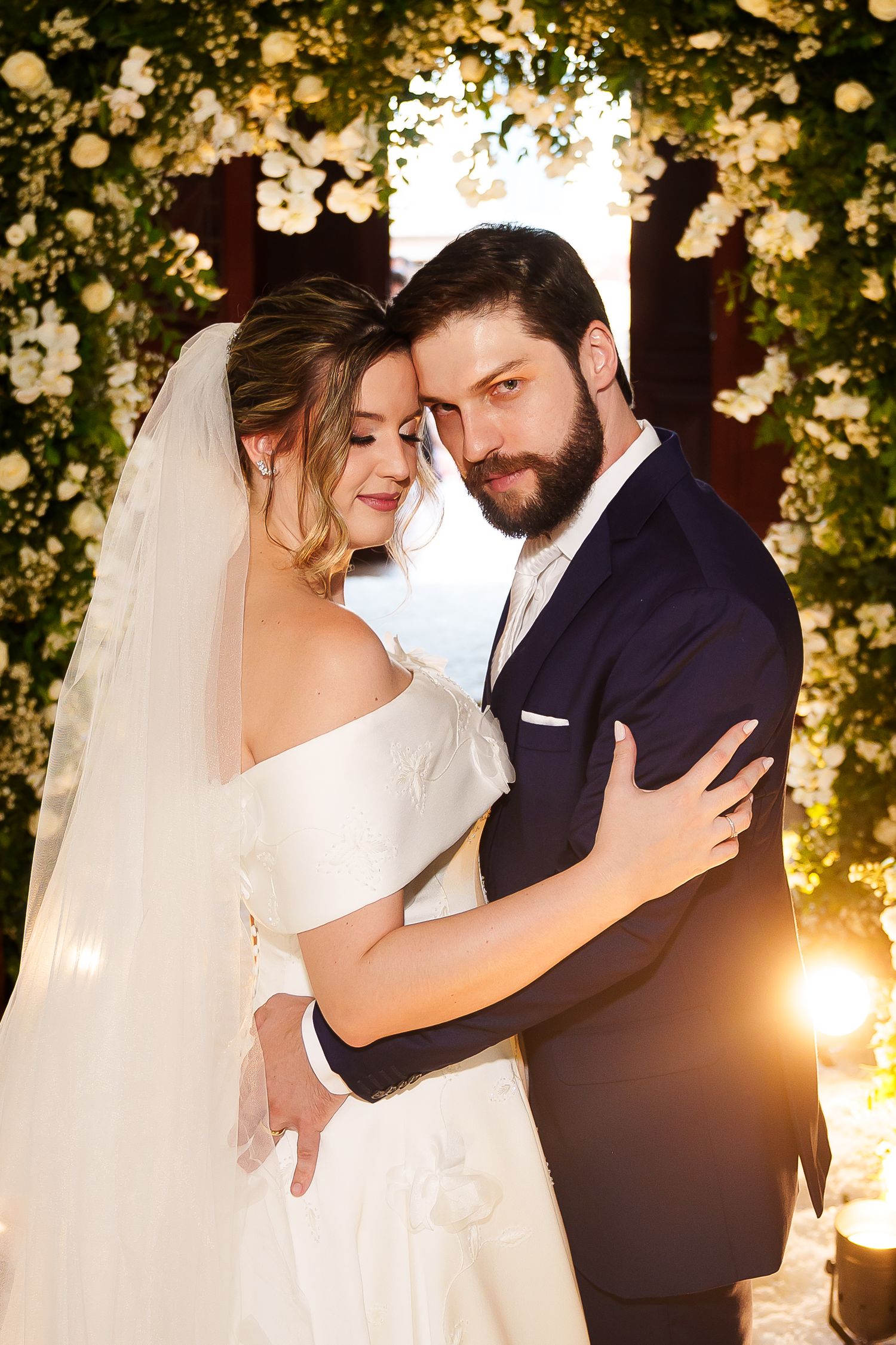 A bride and groom embrace tenderly at their wedding ceremony, surrounded by lush white floral arrangements in warm golden light.