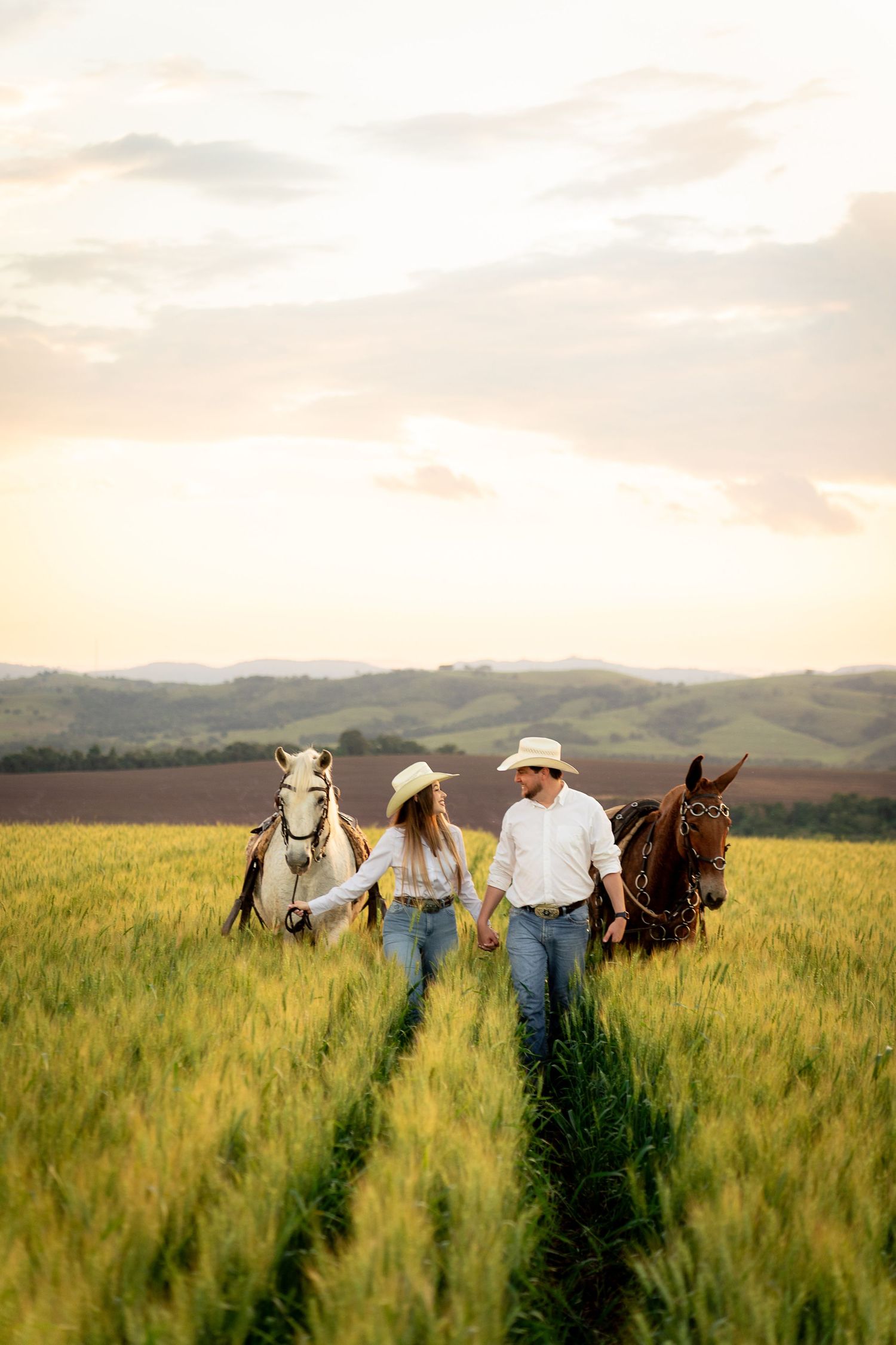 Couple in cowboy hats holding hands walking through green field with two horses at sunset.