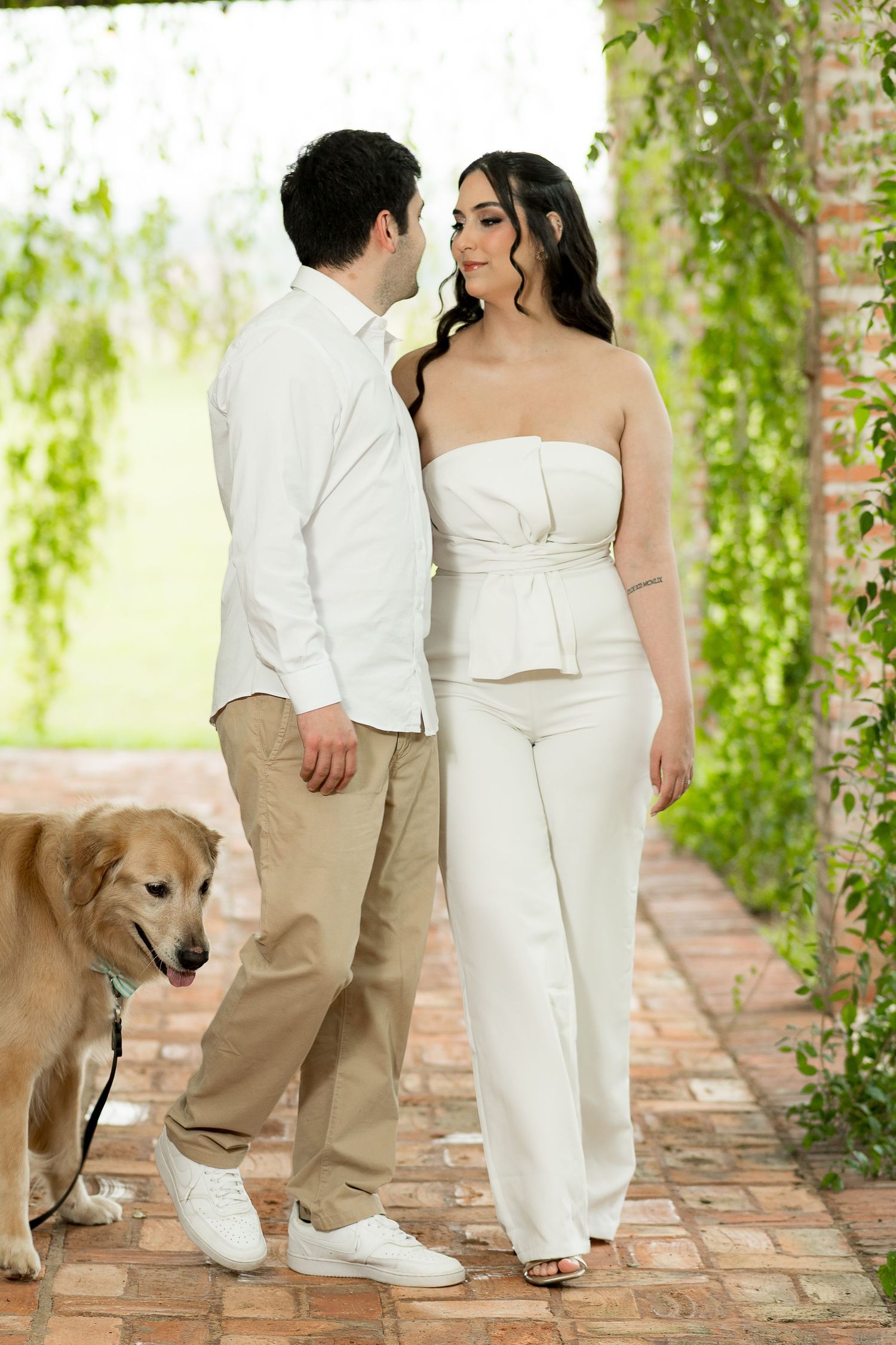 Couple in white outfits walking a golden retriever dog along a brick pathway surrounded by lush green foliage.