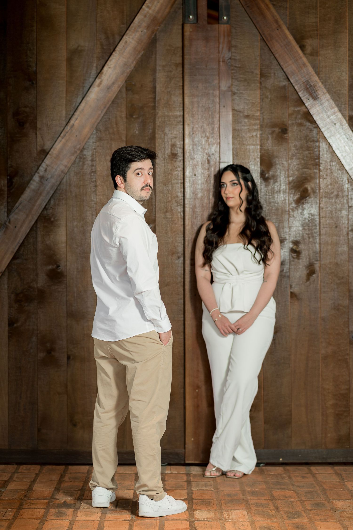 Couple in white outfits posing in front of rustic wooden barn doors, man in chinos and woman in strapless jumpsuit.
