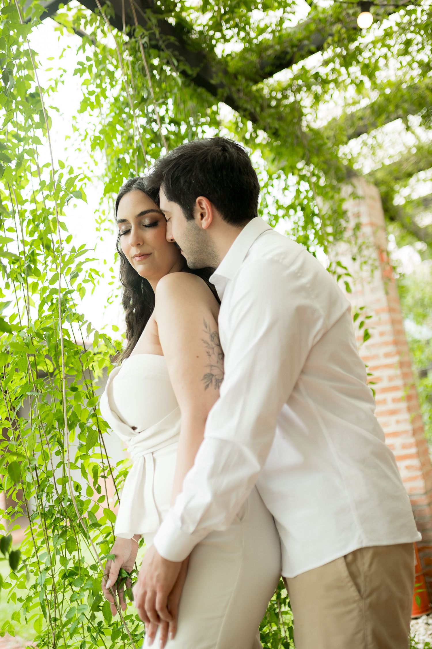 Couple embracing under green vine pergola, woman in white outfit with tattoo, man in white shirt kissing her cheek.