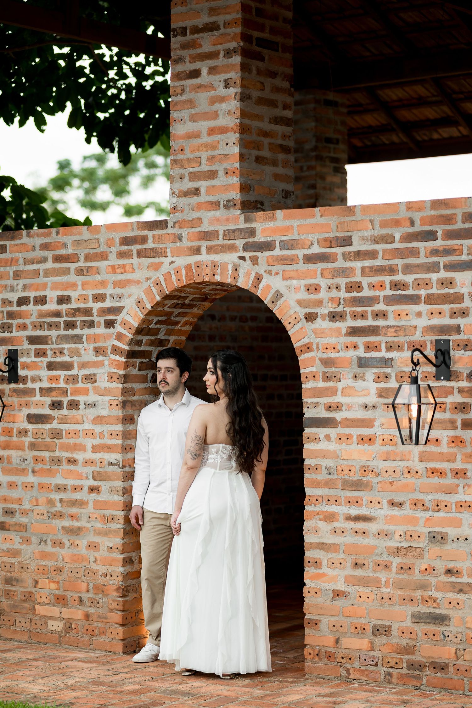 Couple in wedding attire standing in a rustic brick archway, bride in white gown, groom in white shirt and khakis.