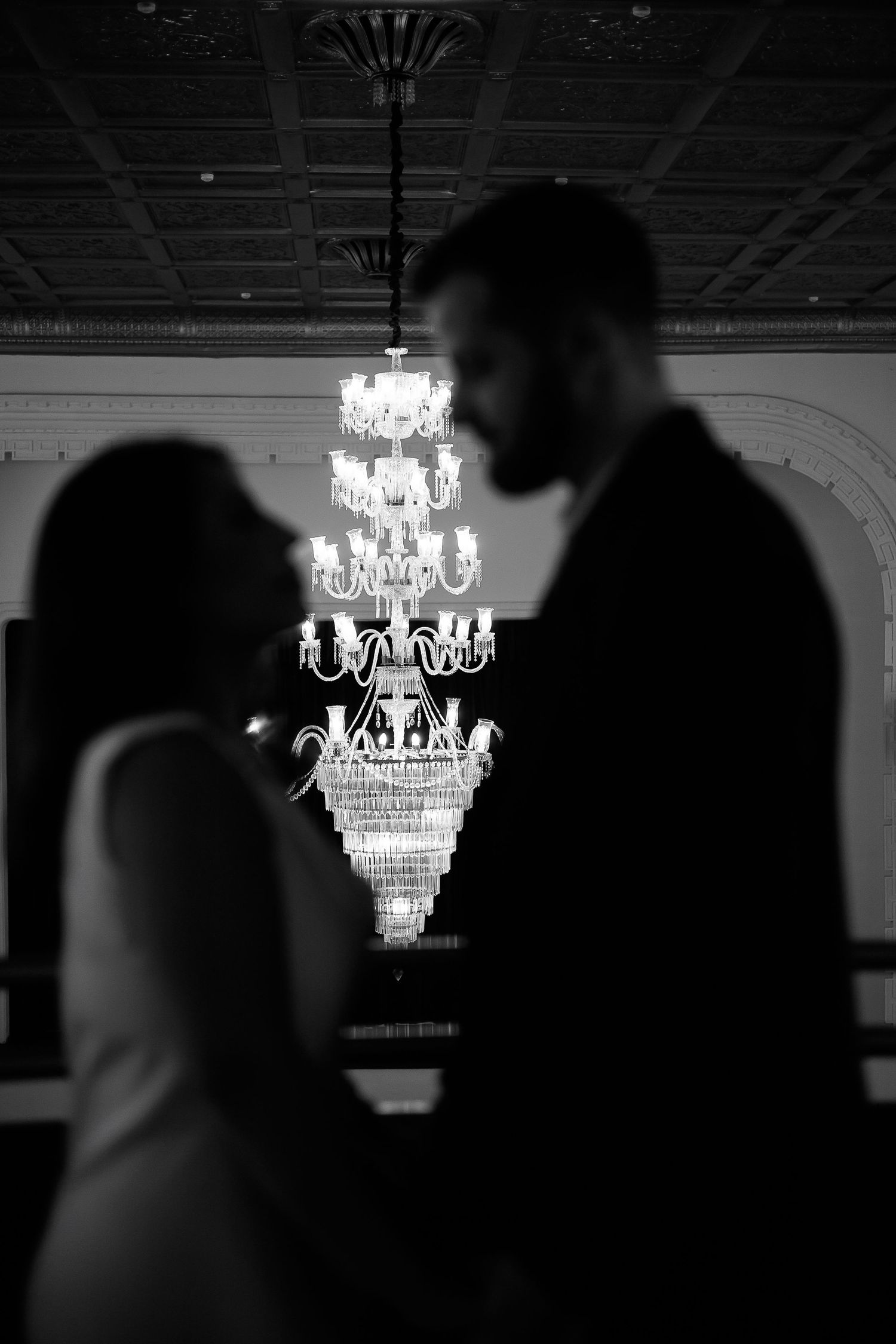 Silhouetted couple sharing romantic moment with elegant crystal chandelier glowing in black and white wedding photo.