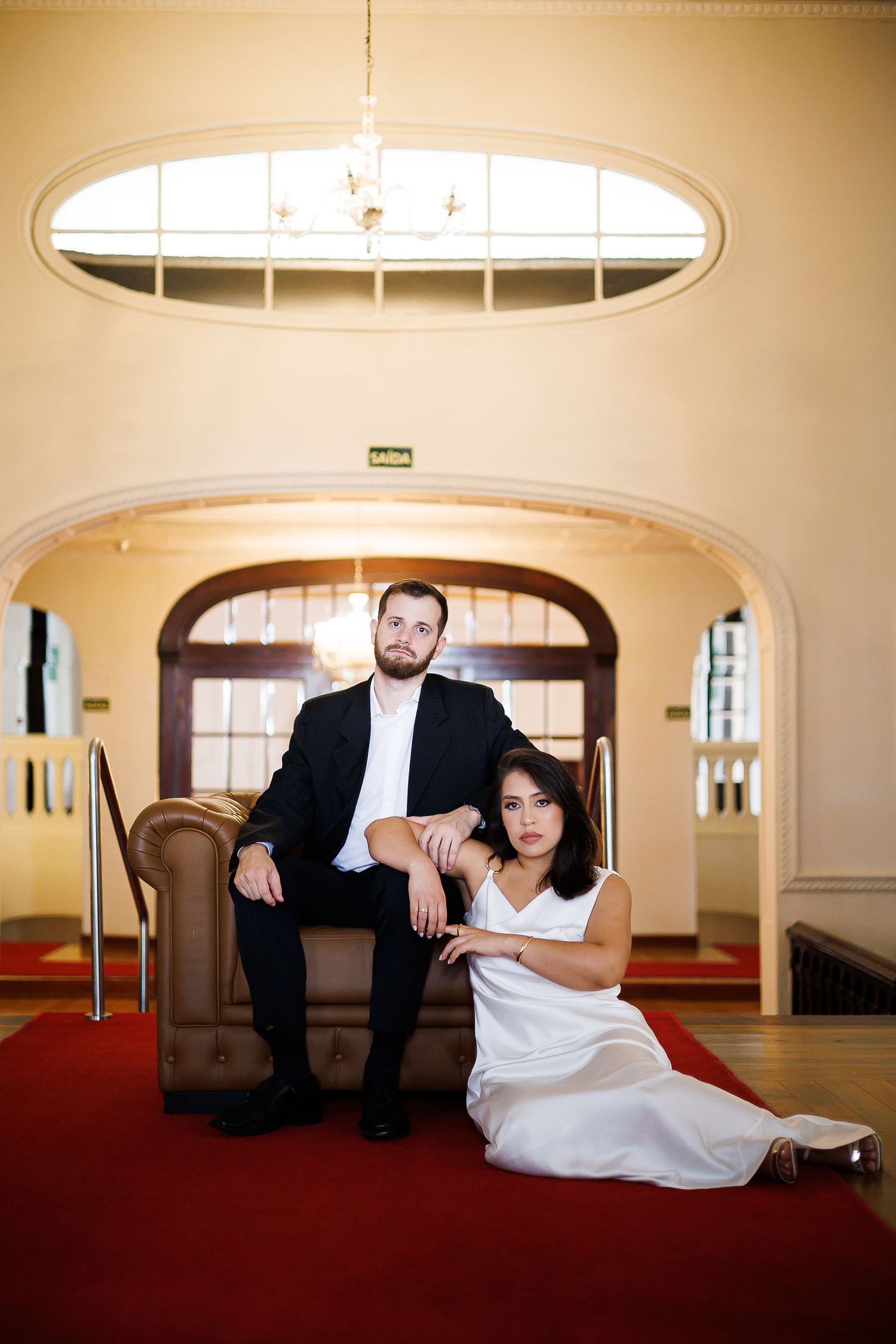 Elegant couple posing in grand hall, man in black suit seated in leather chair, woman in white dress on red carpet floor.