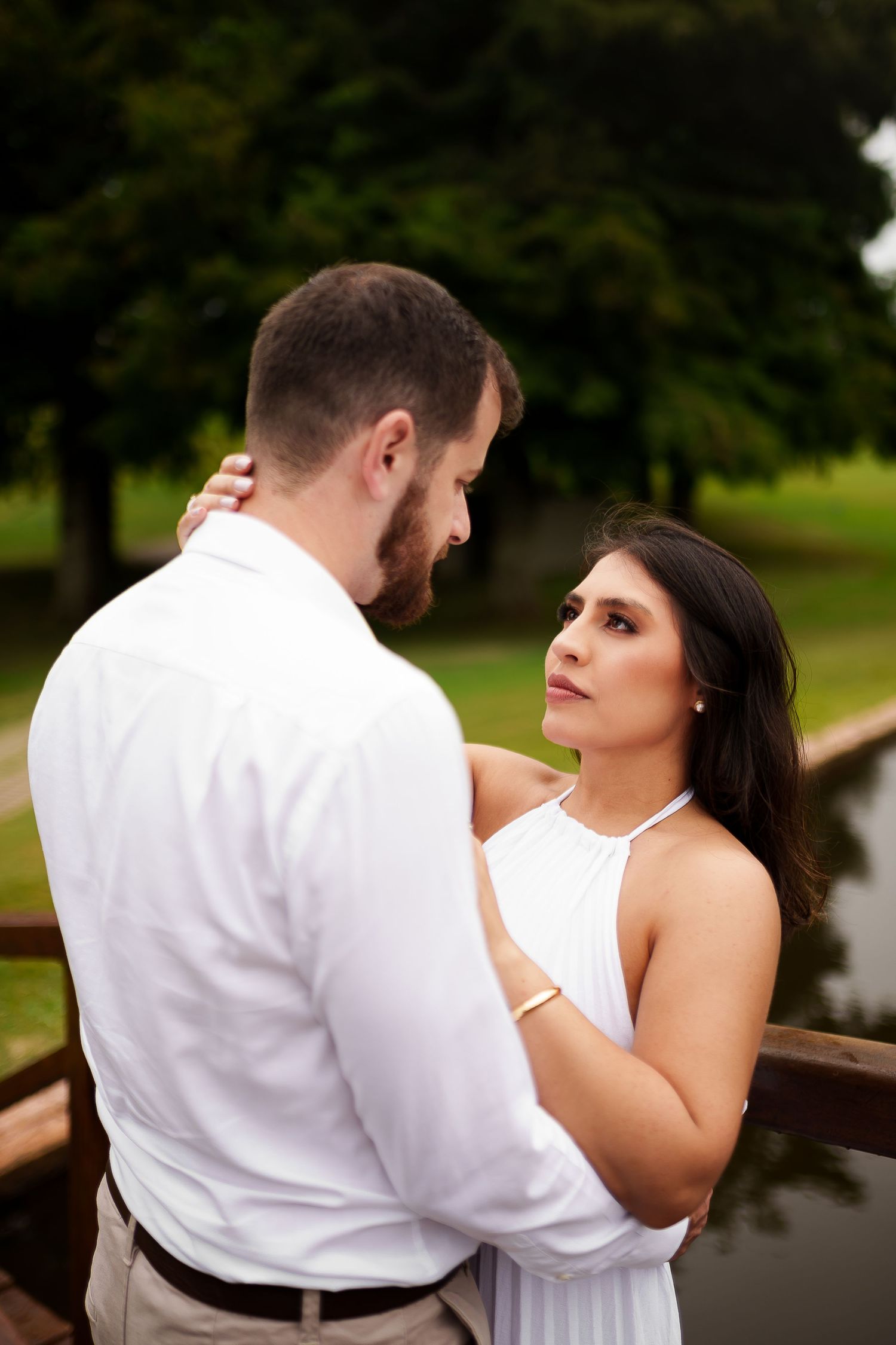 Romantic couple in white outfits sharing intimate moment on bridge surrounded by green trees.