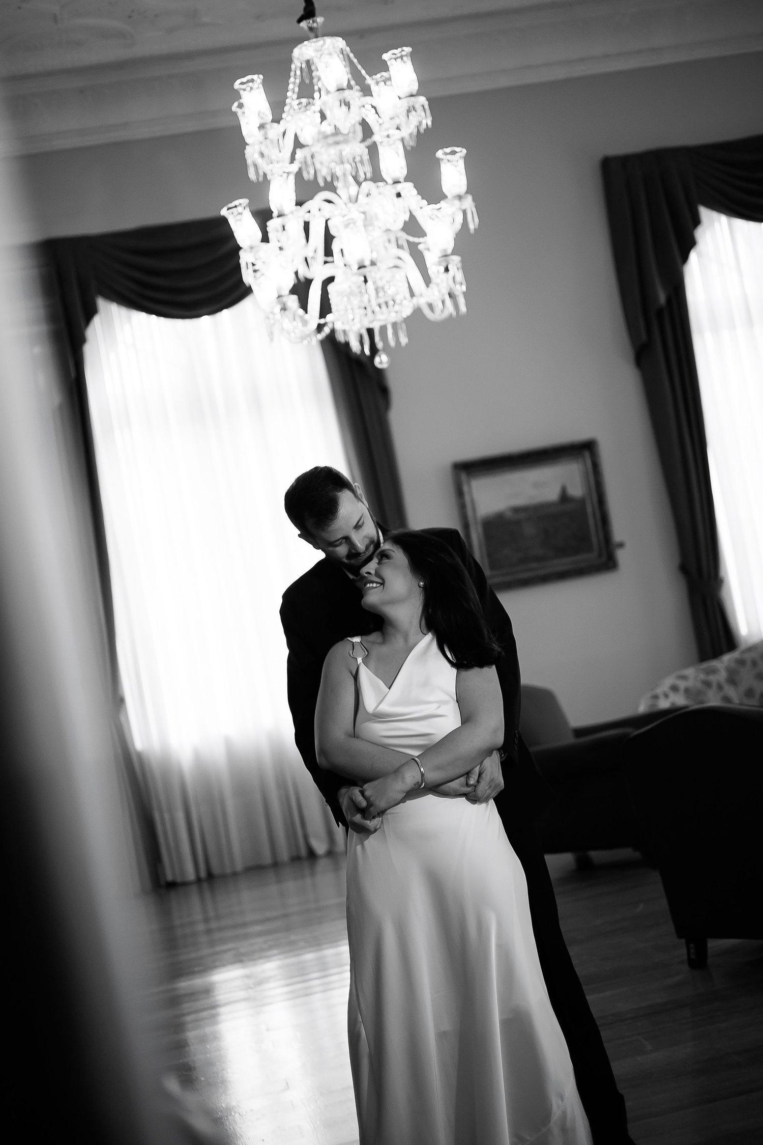 Black and white wedding photo of couple embracing under crystal chandelier in elegant room with tall windows.