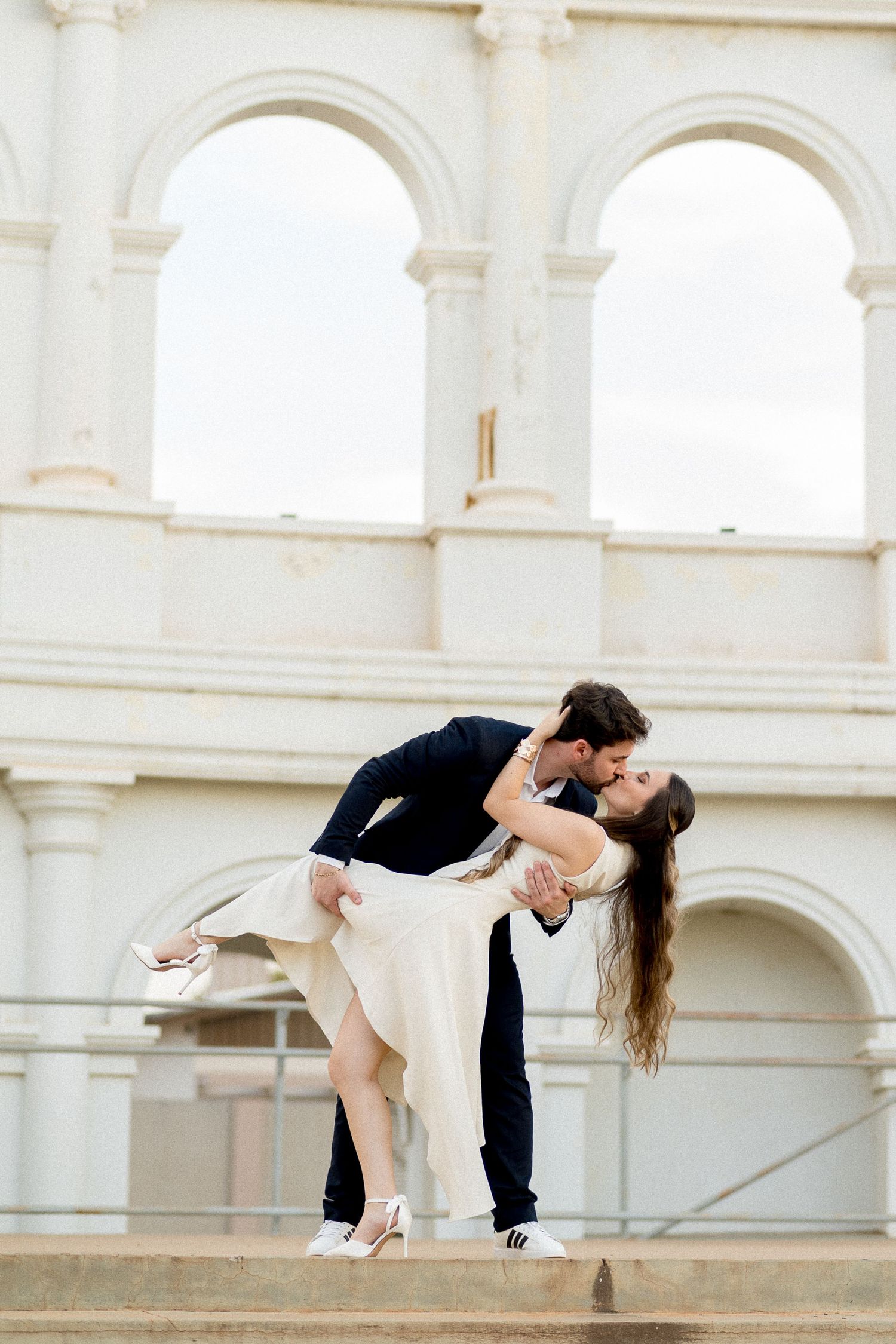 A couple shares a romantic dip kiss on steps before a grand white arched building.