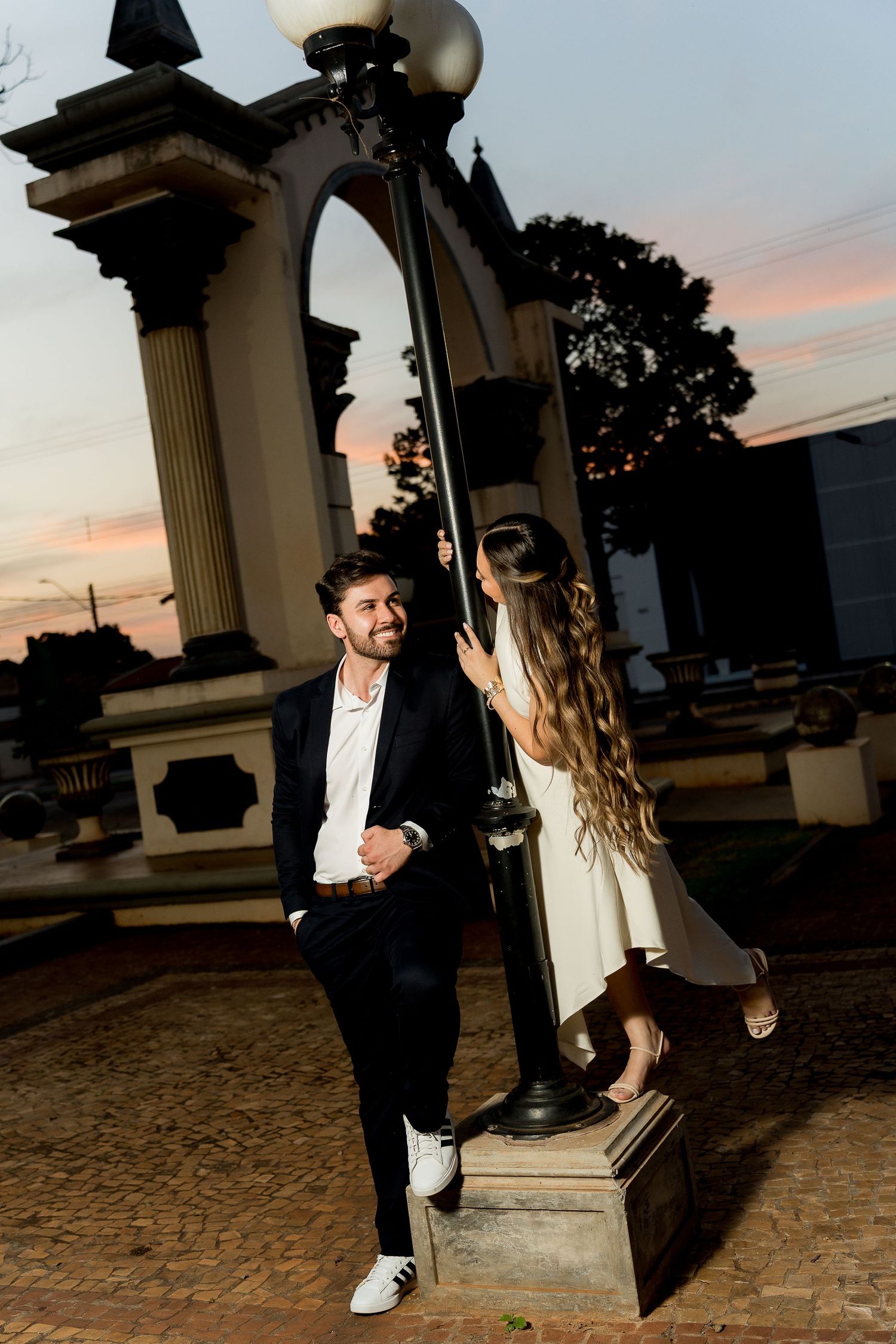 Couple posing by a vintage street lamp at sunset, man in suit smiling at woman in white dress, romantic outdoor setting.
