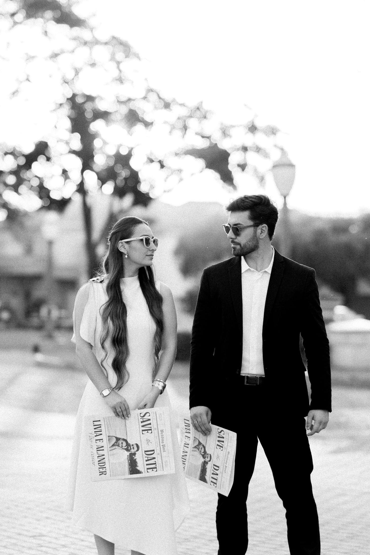Black and white photo of an elegant couple holding Save the Date cards outdoors, wearing sunglasses and formal attire.