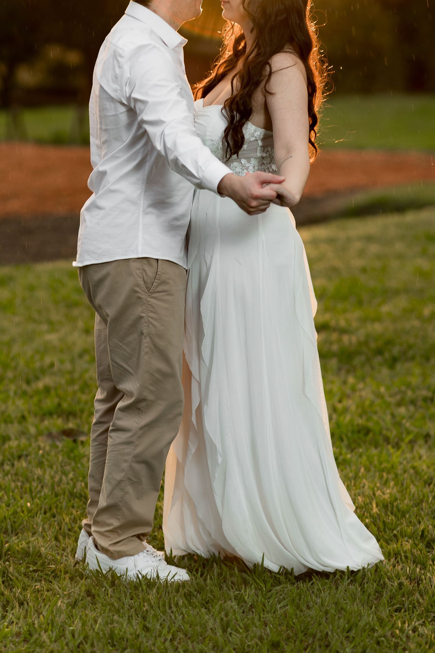 Couple embracing outdoors at sunset, man in white shirt and khakis, woman in flowing white gown on green grass.