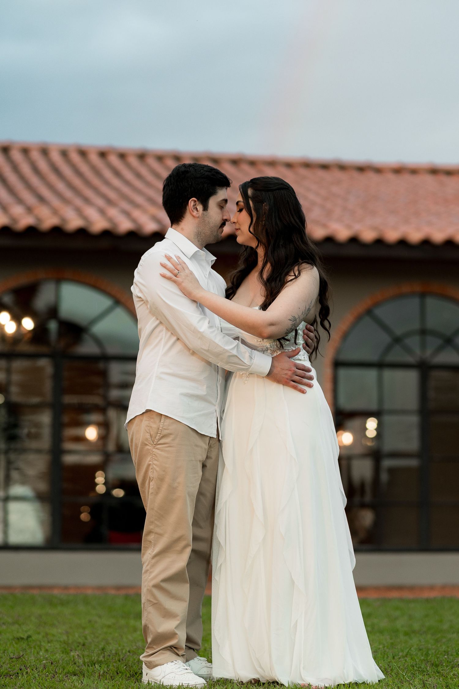 Couple sharing a romantic moment in front of a rustic venue with a rainbow in the background.