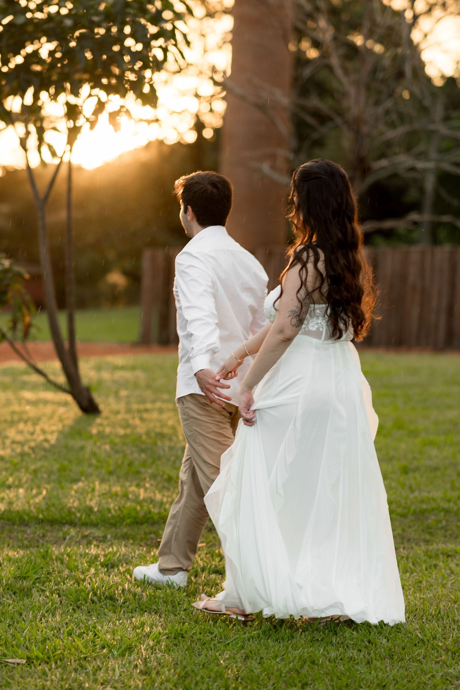 Couple holding hands walking through garden at golden sunset, woman in flowing white gown, man in white shirt and khaki pants.