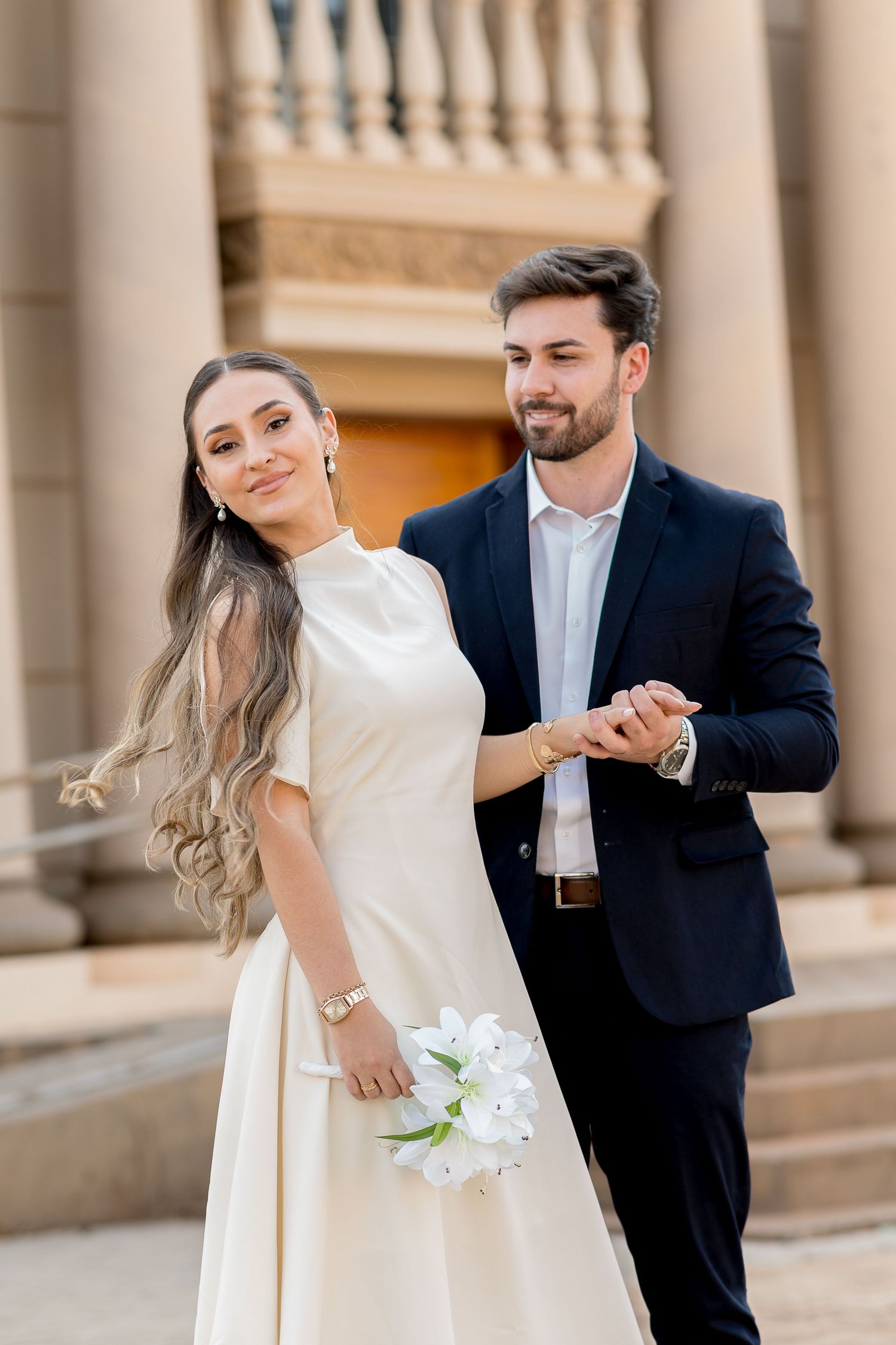 Elegant bride in white gown holds flowers while groom in navy suit holds her hand outside grand building.