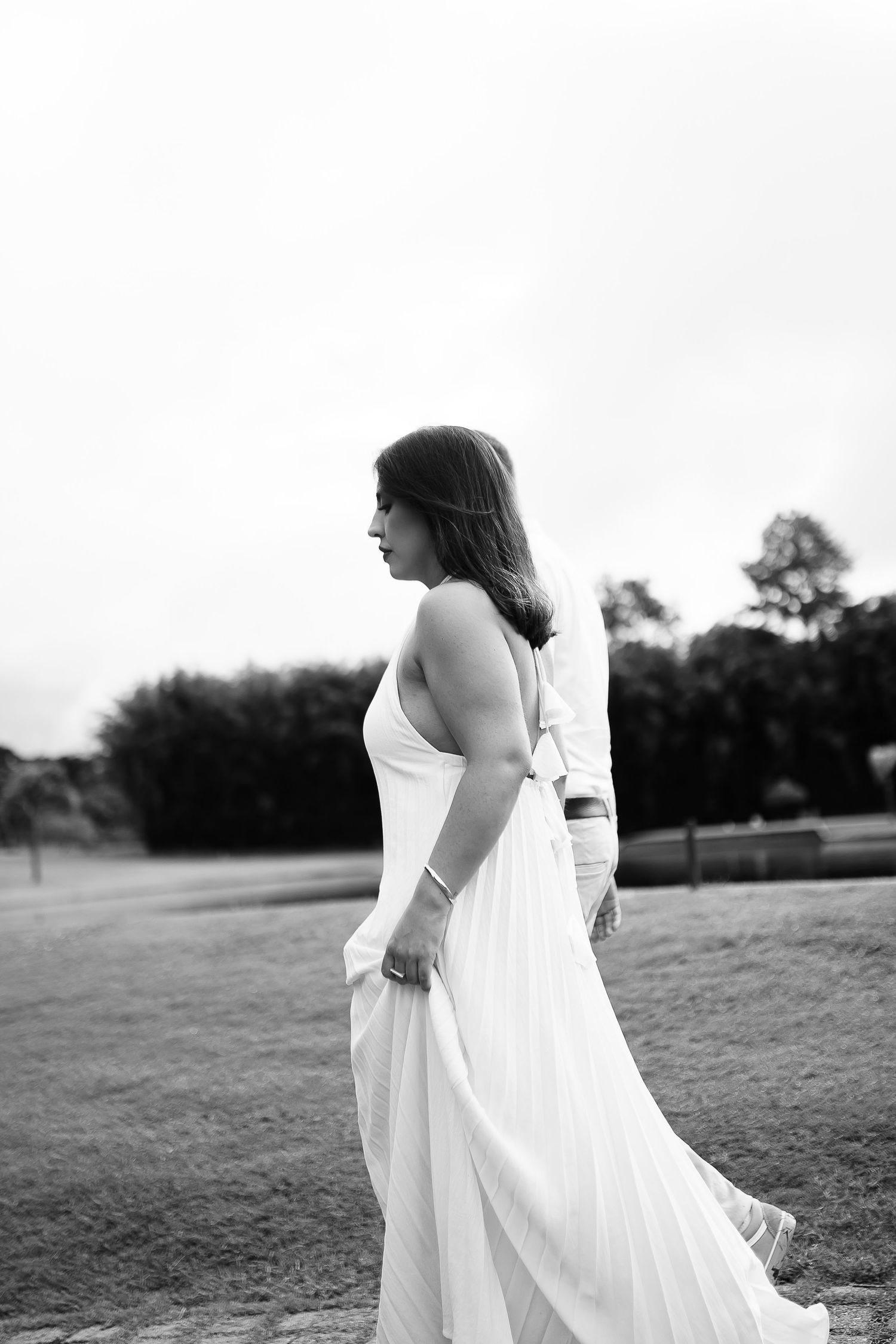 Black and white photo of a woman in a flowing white halter gown walking gracefully outdoors on a grassy field.