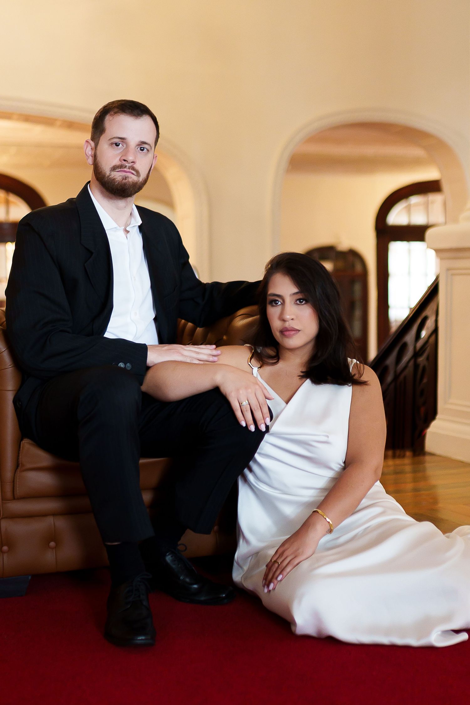 Elegant couple in formal attire posing in a grand interior, man in black suit seated behind woman in white dress.