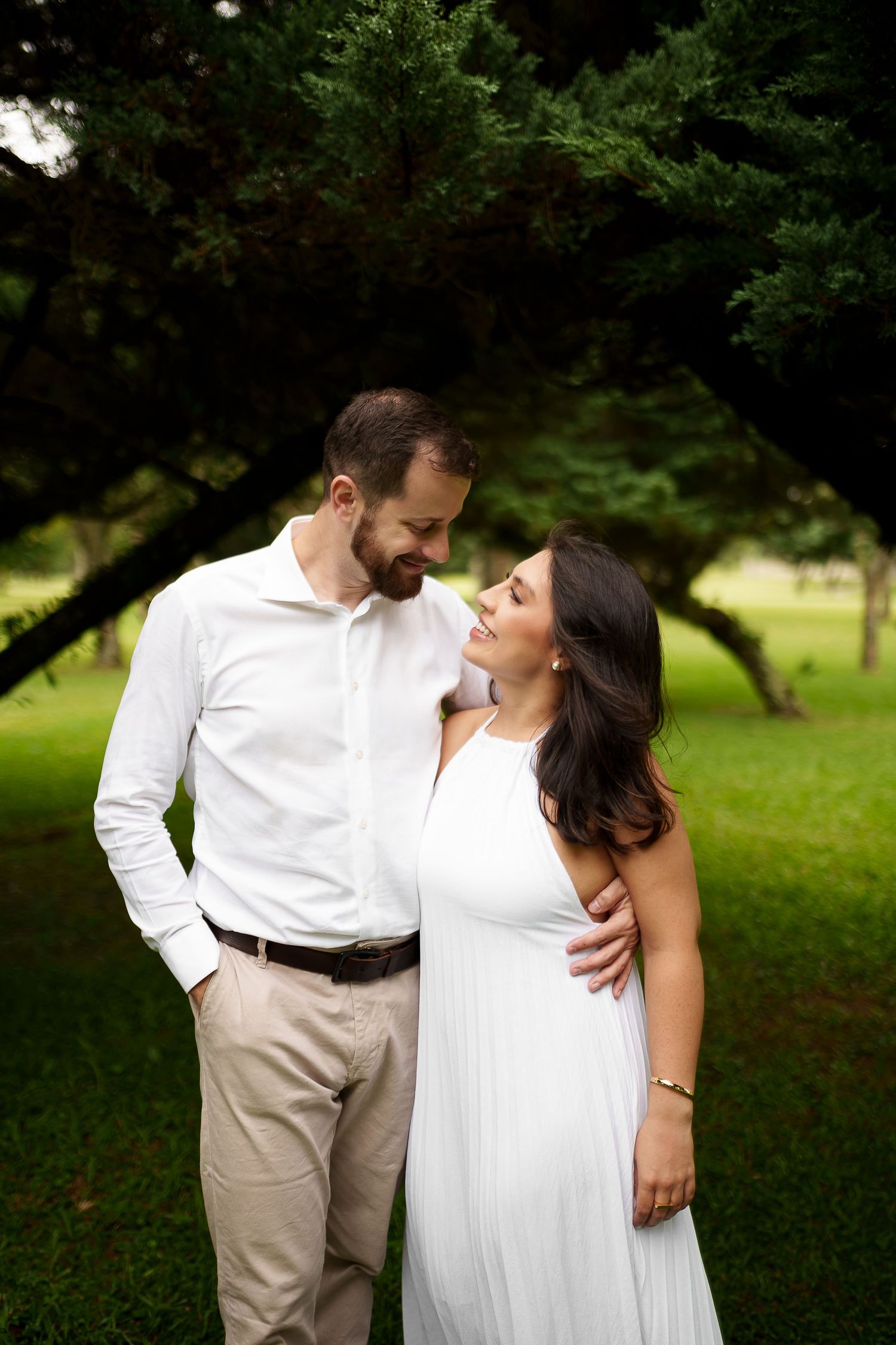 A couple in white outfits share a tender moment outdoors, smiling at each other beneath lush green trees.