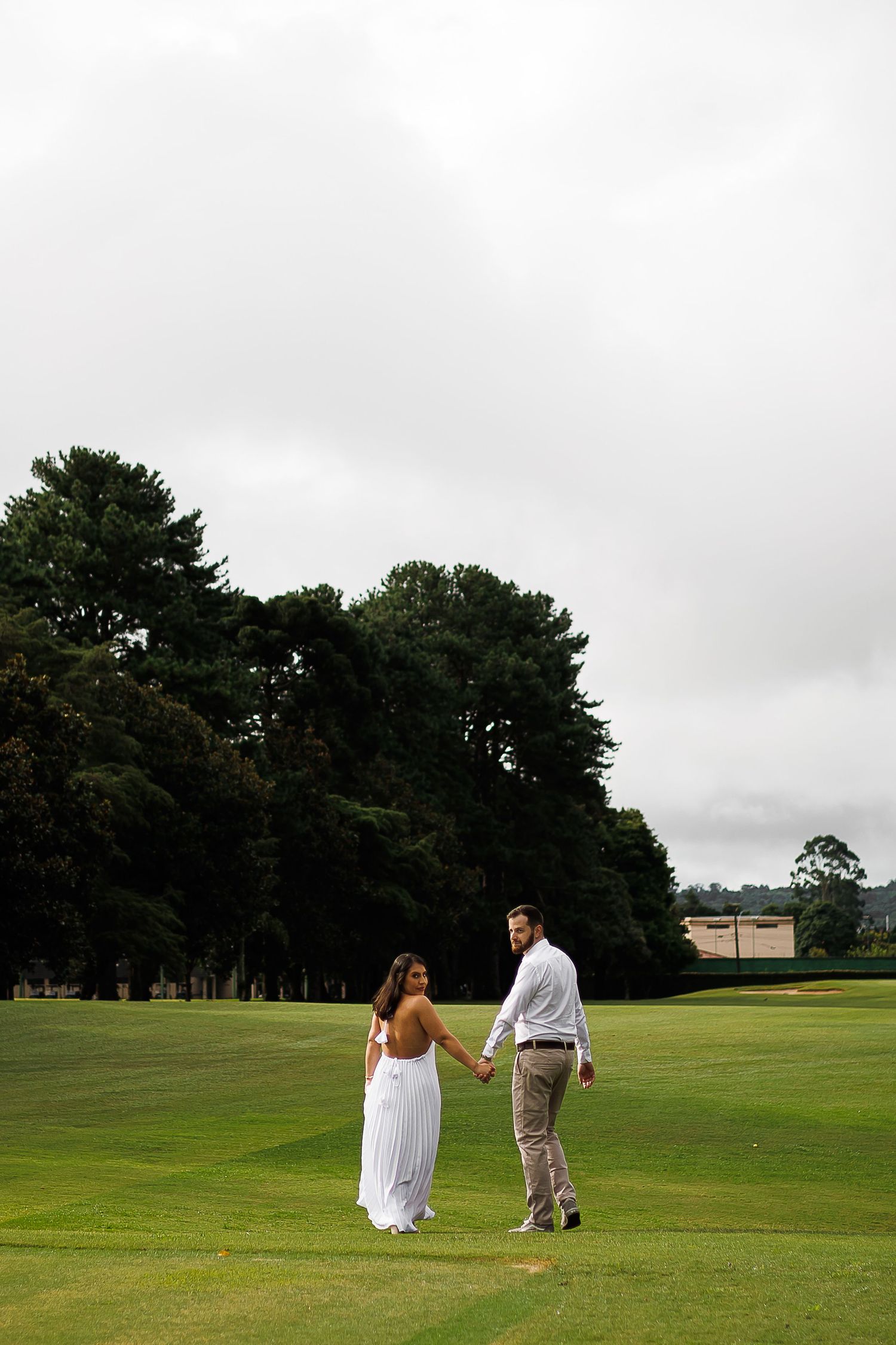 Couple holding hands on lush green golf course, woman in white backless gown, man in casual attire, tall trees behind.