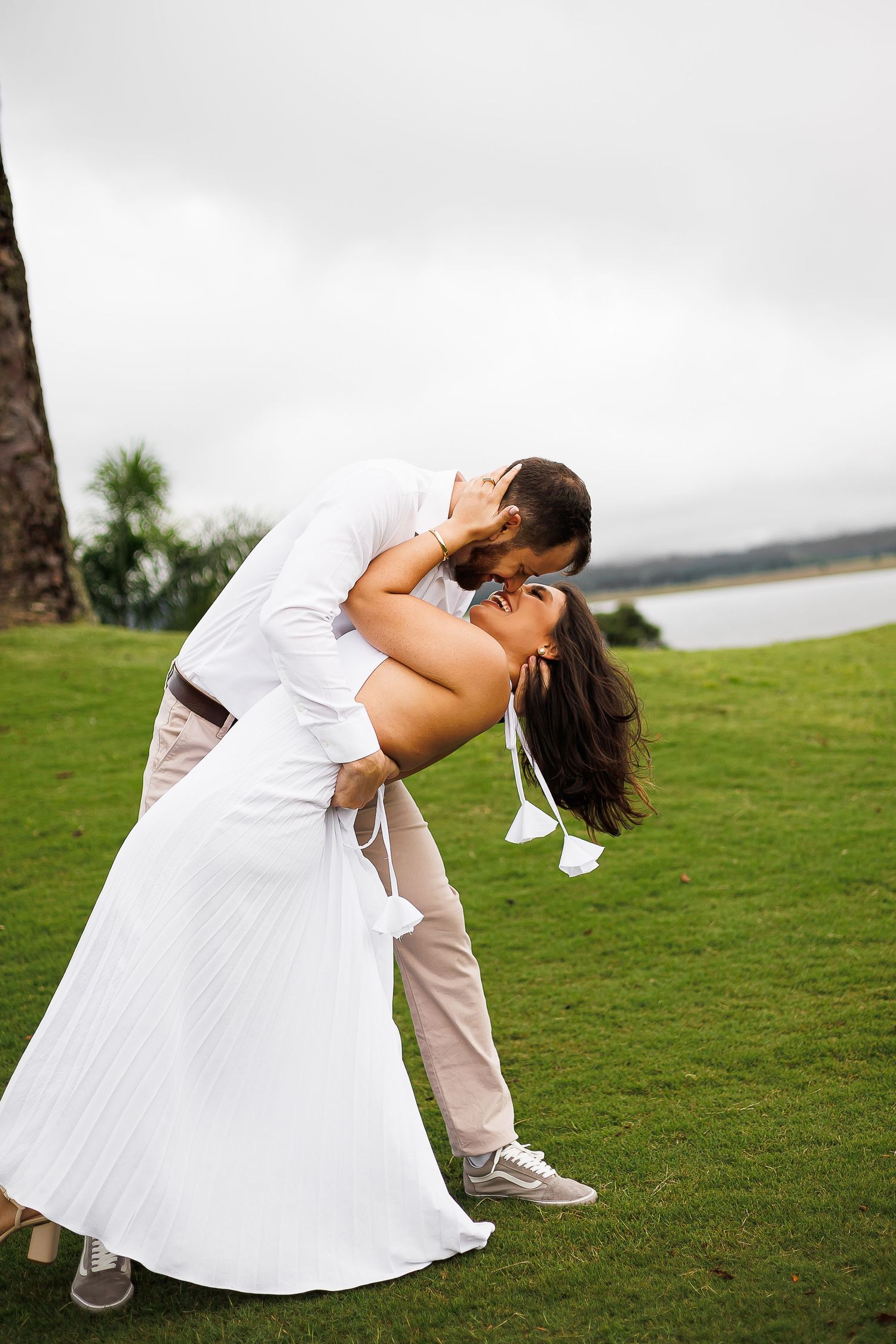 A couple shares a romantic dip kiss on a lush green lawn near a waterfront on their wedding day.