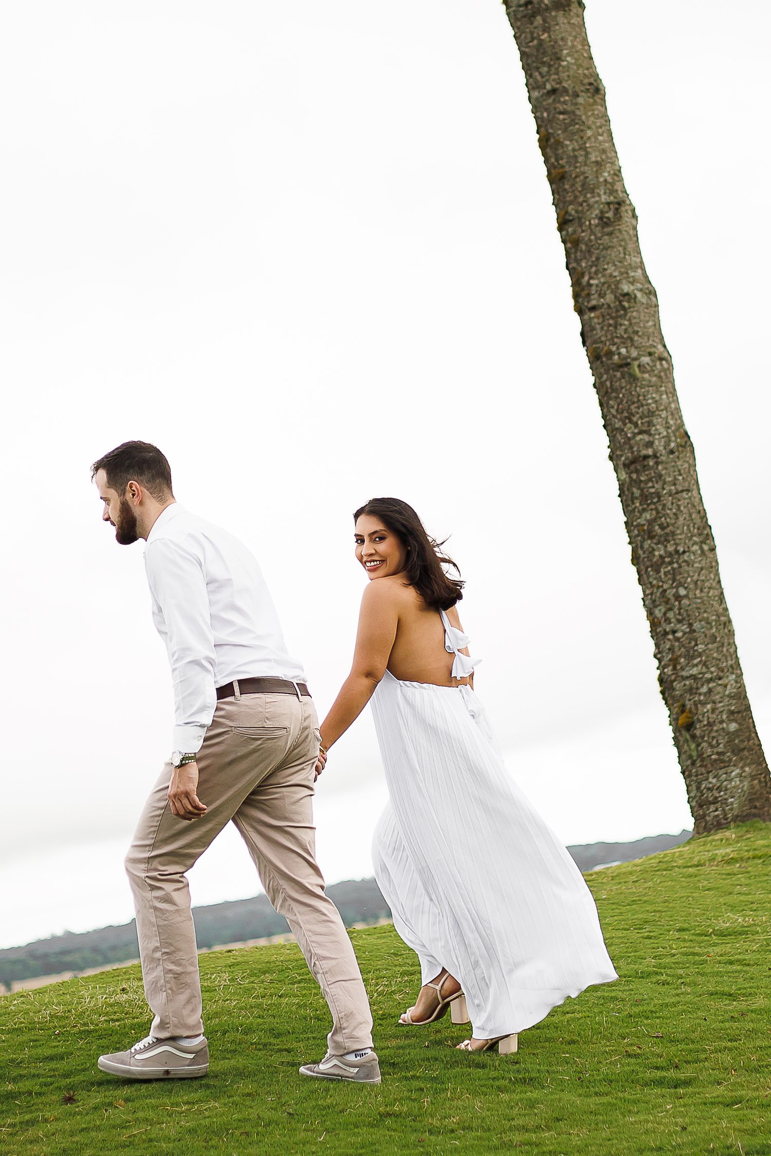 Couple in white and beige outfits holding hands while walking on green grass near a tall tree.