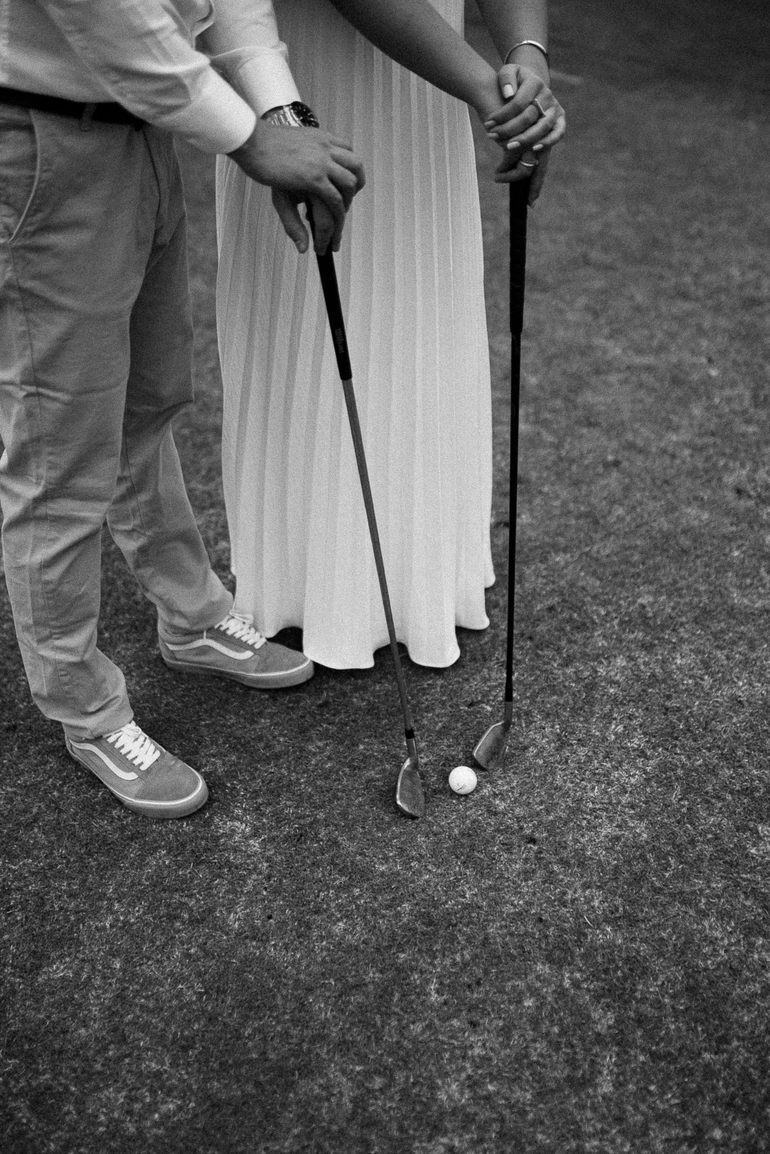Black and white photo of a couple holding golf clubs over a golf ball on a course, wearing casual attire.
