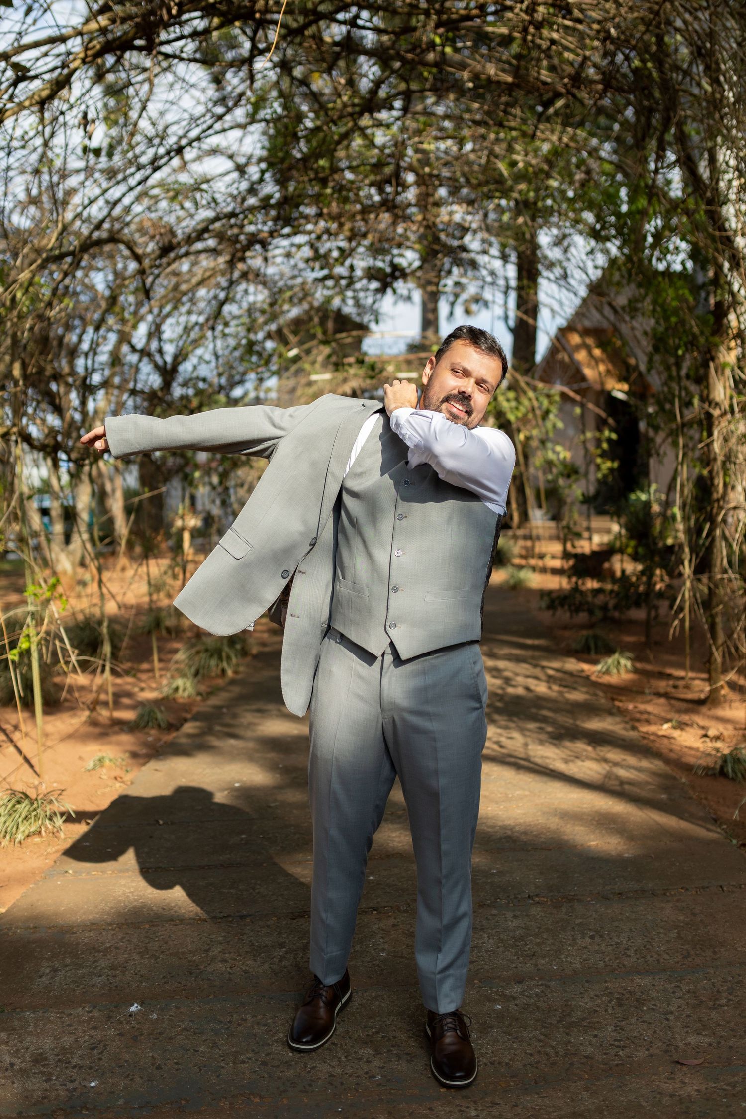 Man in gray suit playfully swinging his jacket outdoors in a garden setting.
