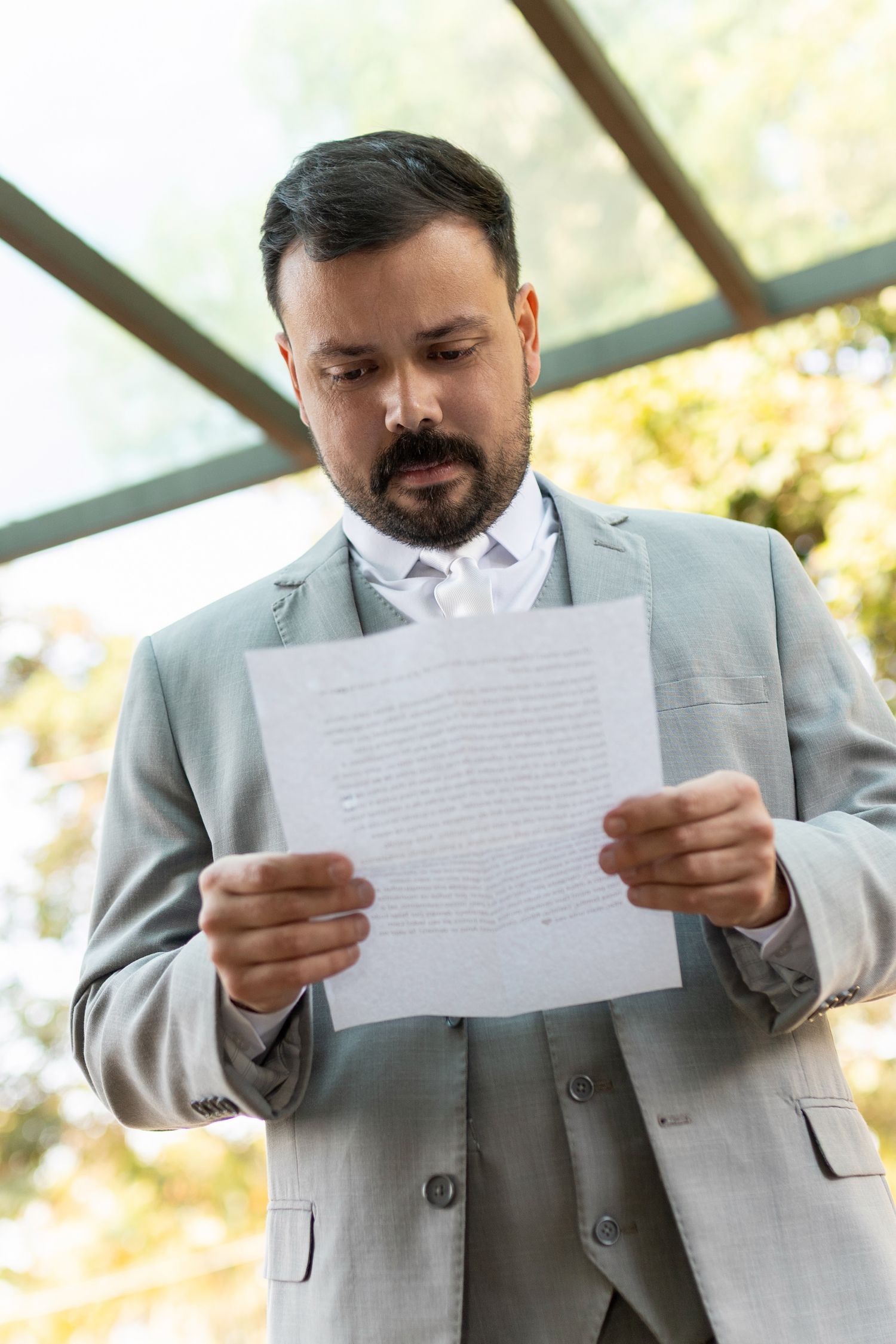 Businessman in gray suit reading document outdoors near glass building.