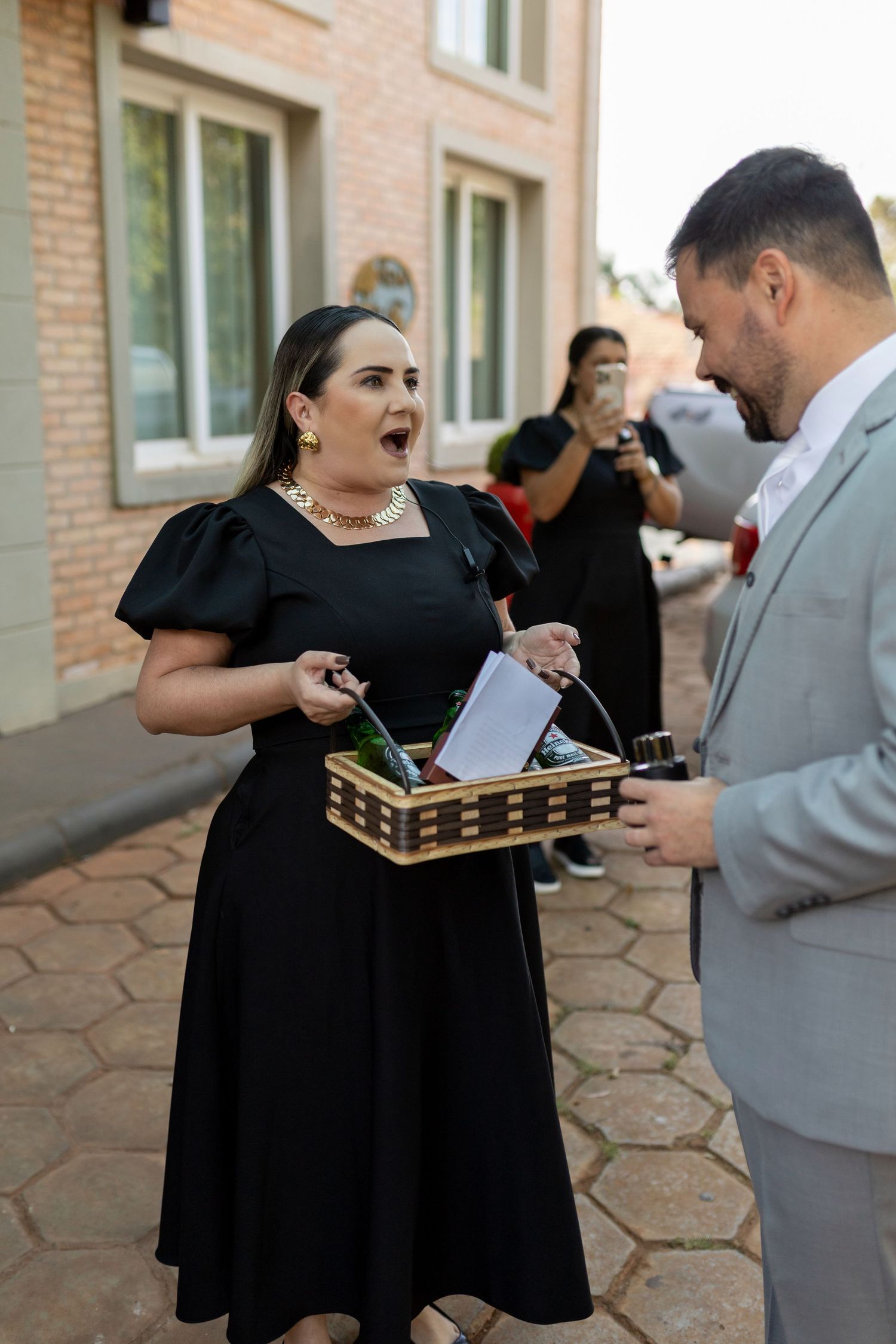 Woman in black dress holds wicker basket while talking to man in gray suit outdoors near brick building.