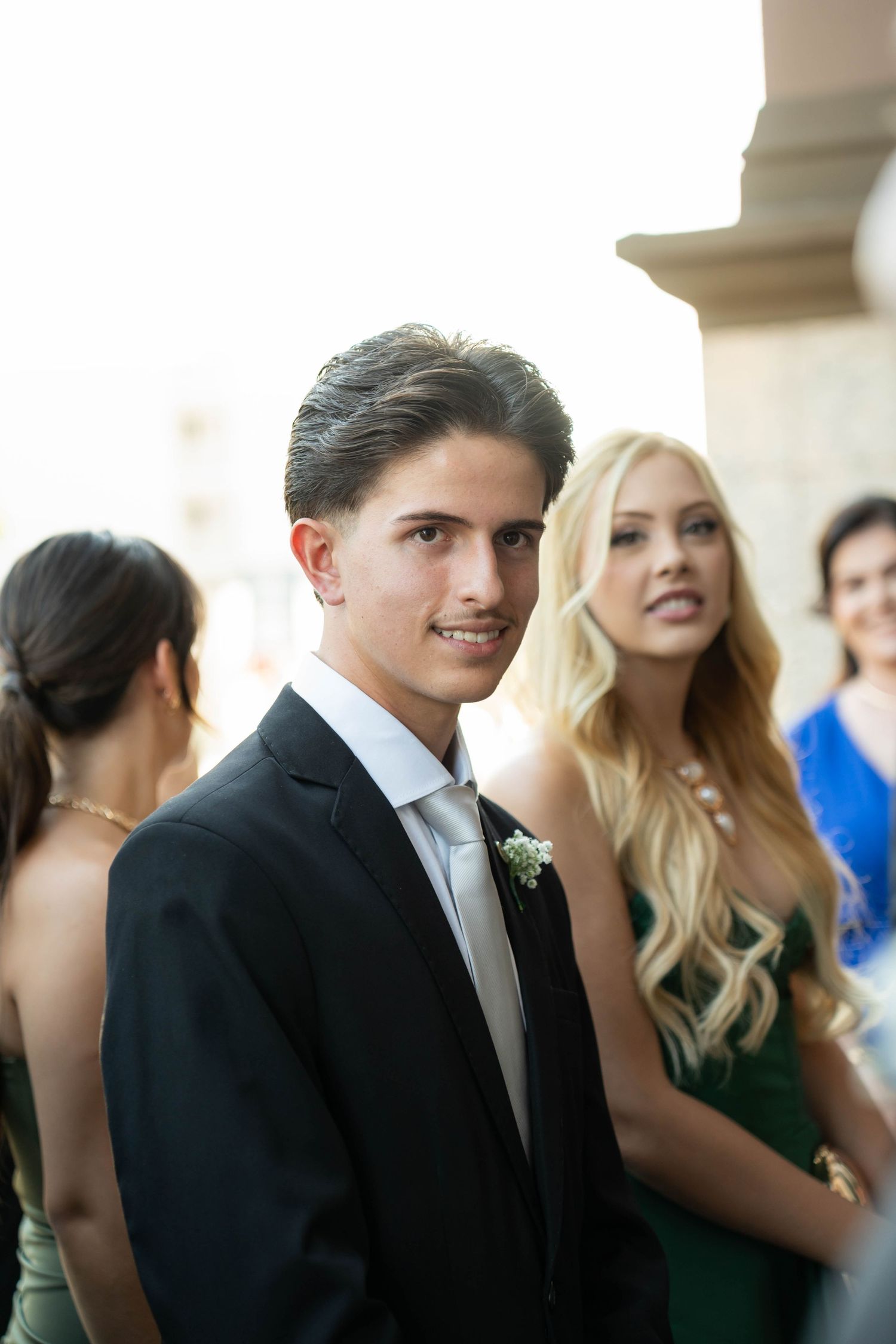 Young man in black suit with boutonniere smiles at camera, blonde woman in green dress stands behind him.