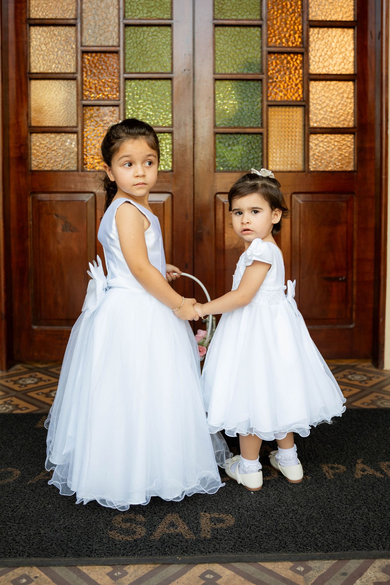Two young flower girls in white dresses holding a basket stand before ornate wooden doors with stained glass panels.