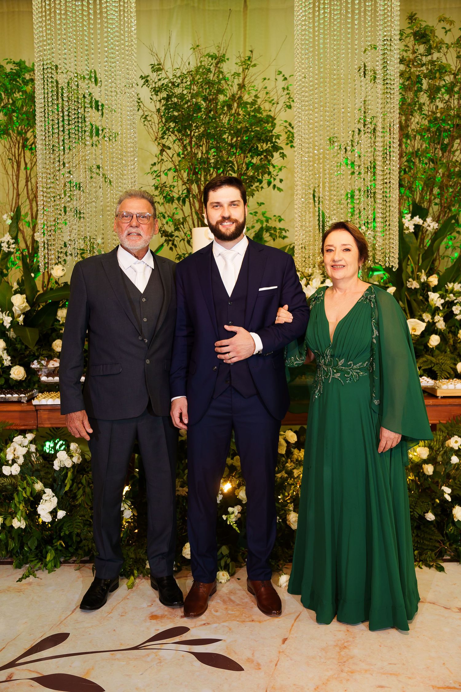 Three elegantly dressed people pose together at a formal event, with a man in a navy suit flanked by two others.