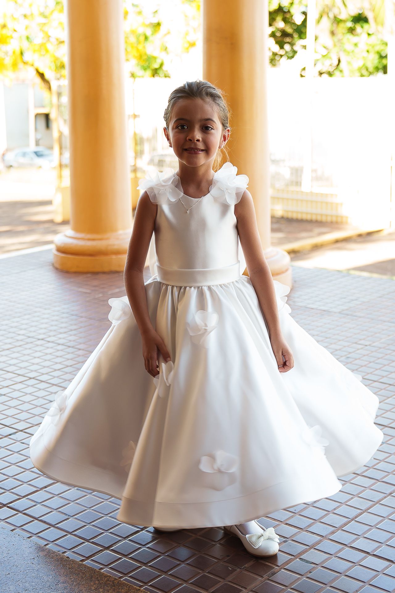 Young girl in elegant white first communion dress with bow details, standing in a columned outdoor setting.