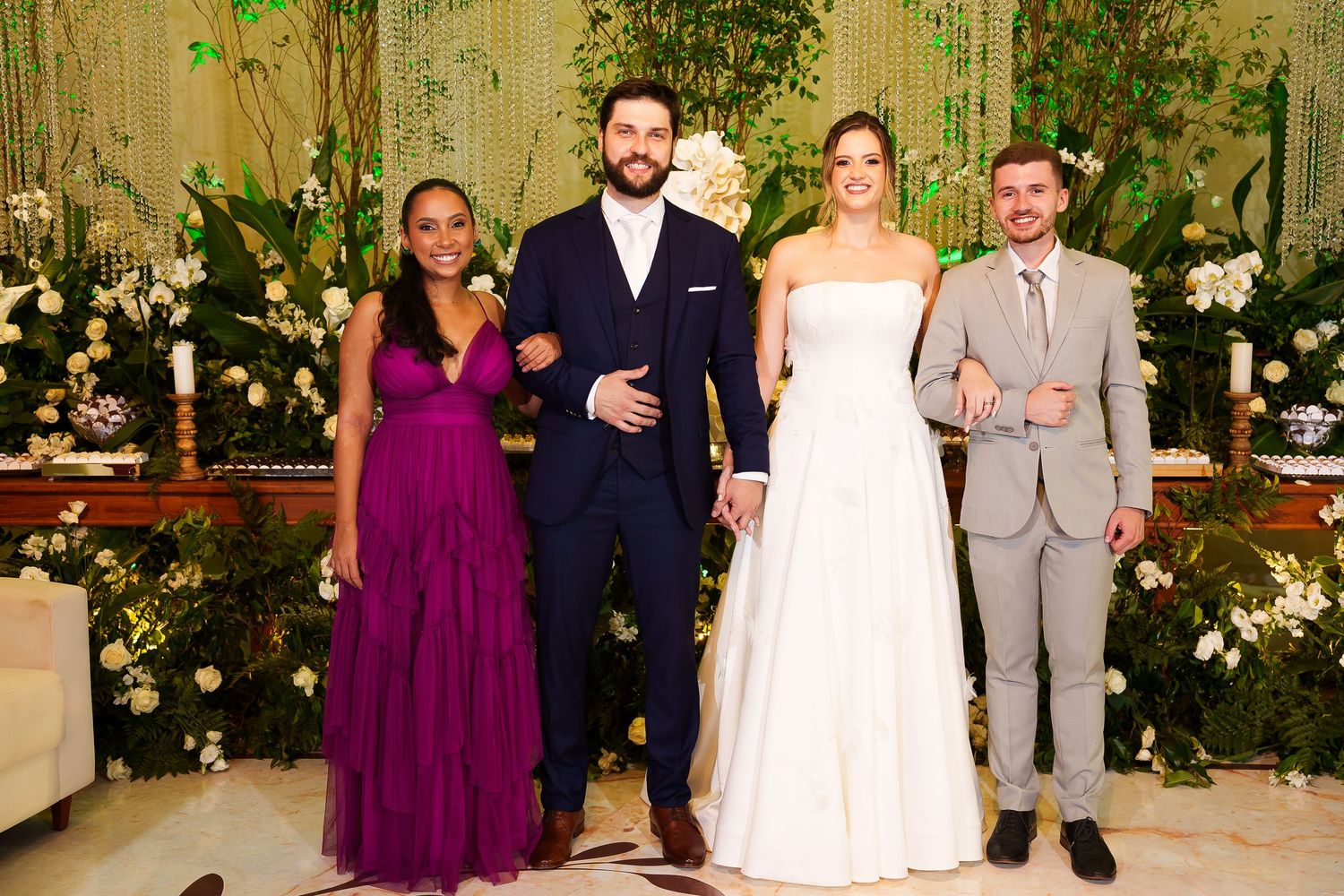 Four elegantly dressed wedding guests pose together in front of a lush floral backdrop at a formal ceremony.