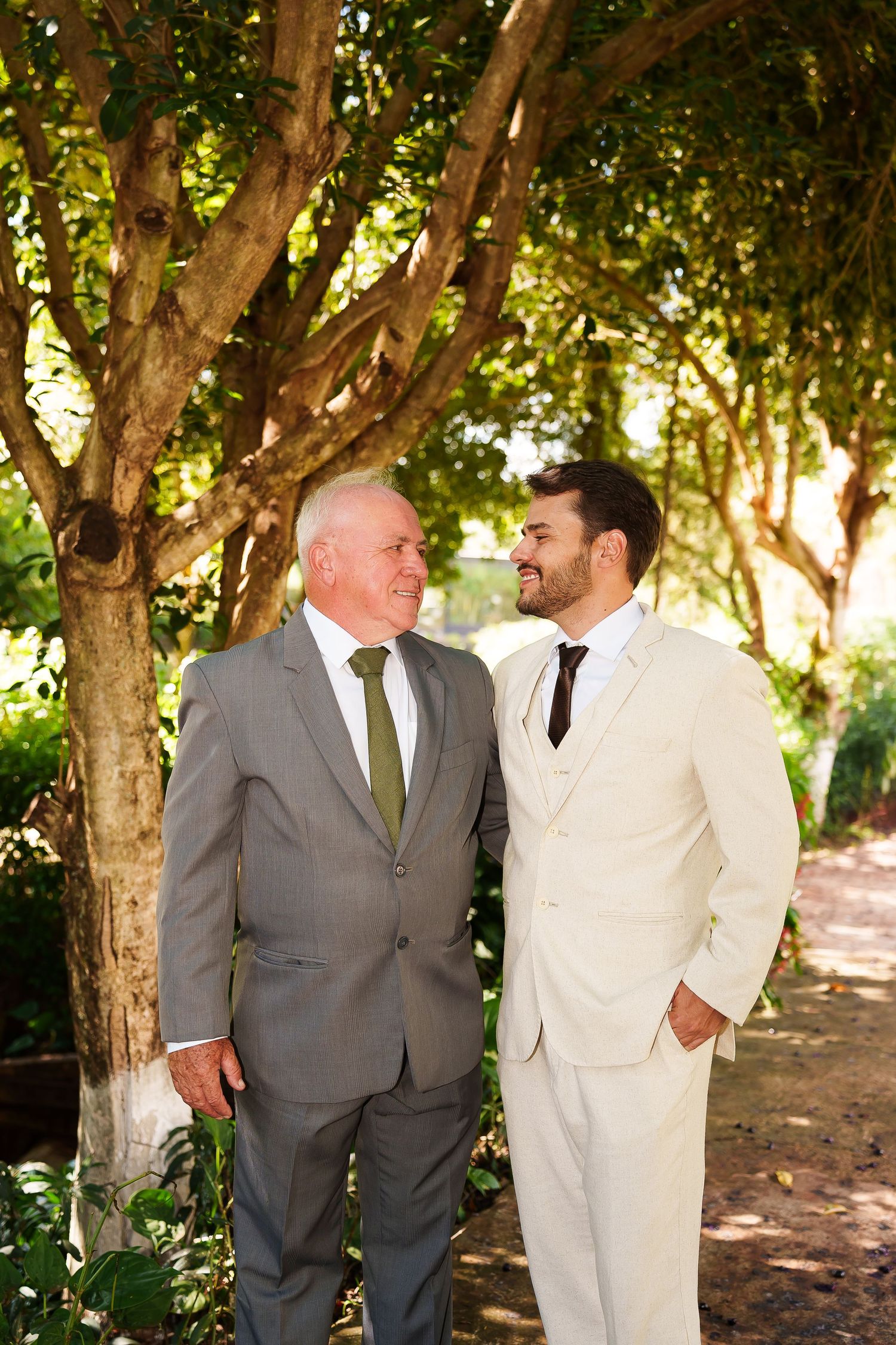 Two men in suits smiling at each other outdoors among green trees, likely at a wedding celebration.