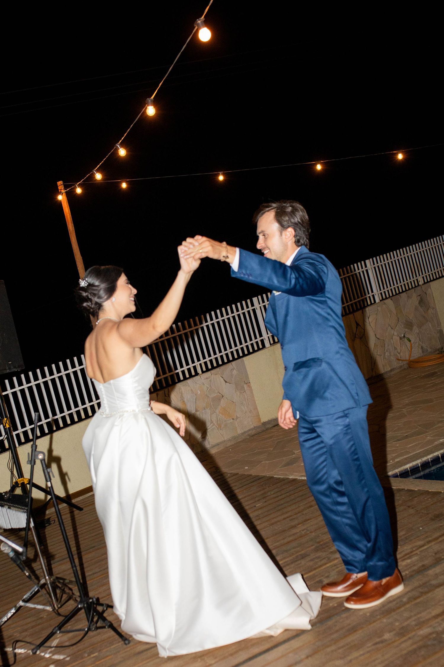 Bride and groom share their first dance outdoors at night under string lights, bride in white gown, groom in blue suit.