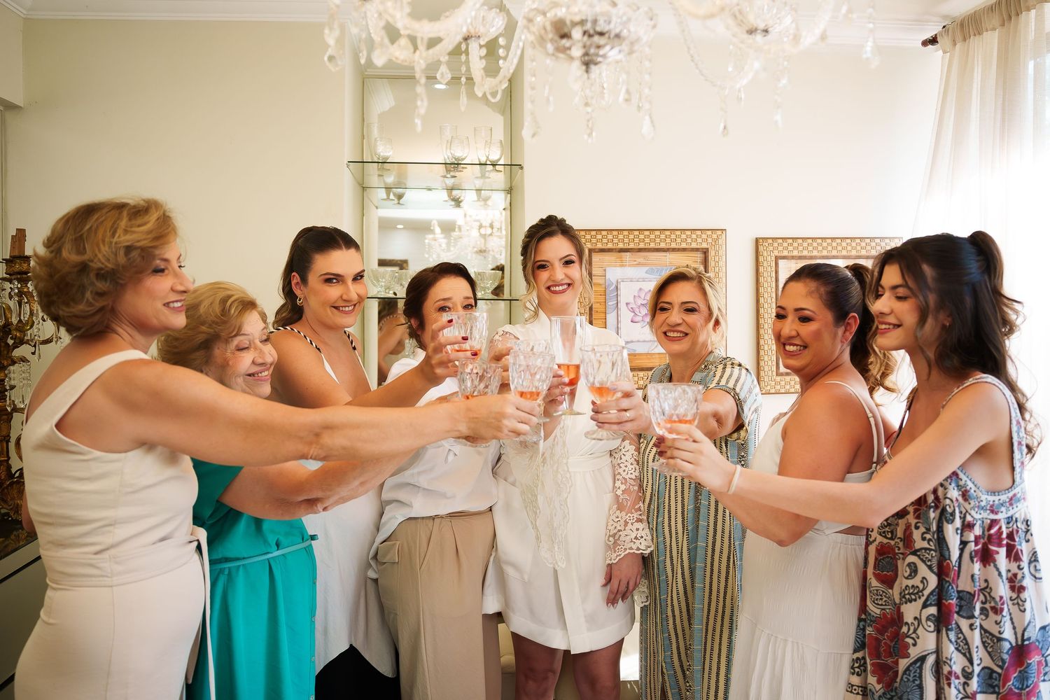 A bride in white toasts champagne with her bridesmaids and family in an elegant room with a chandelier.