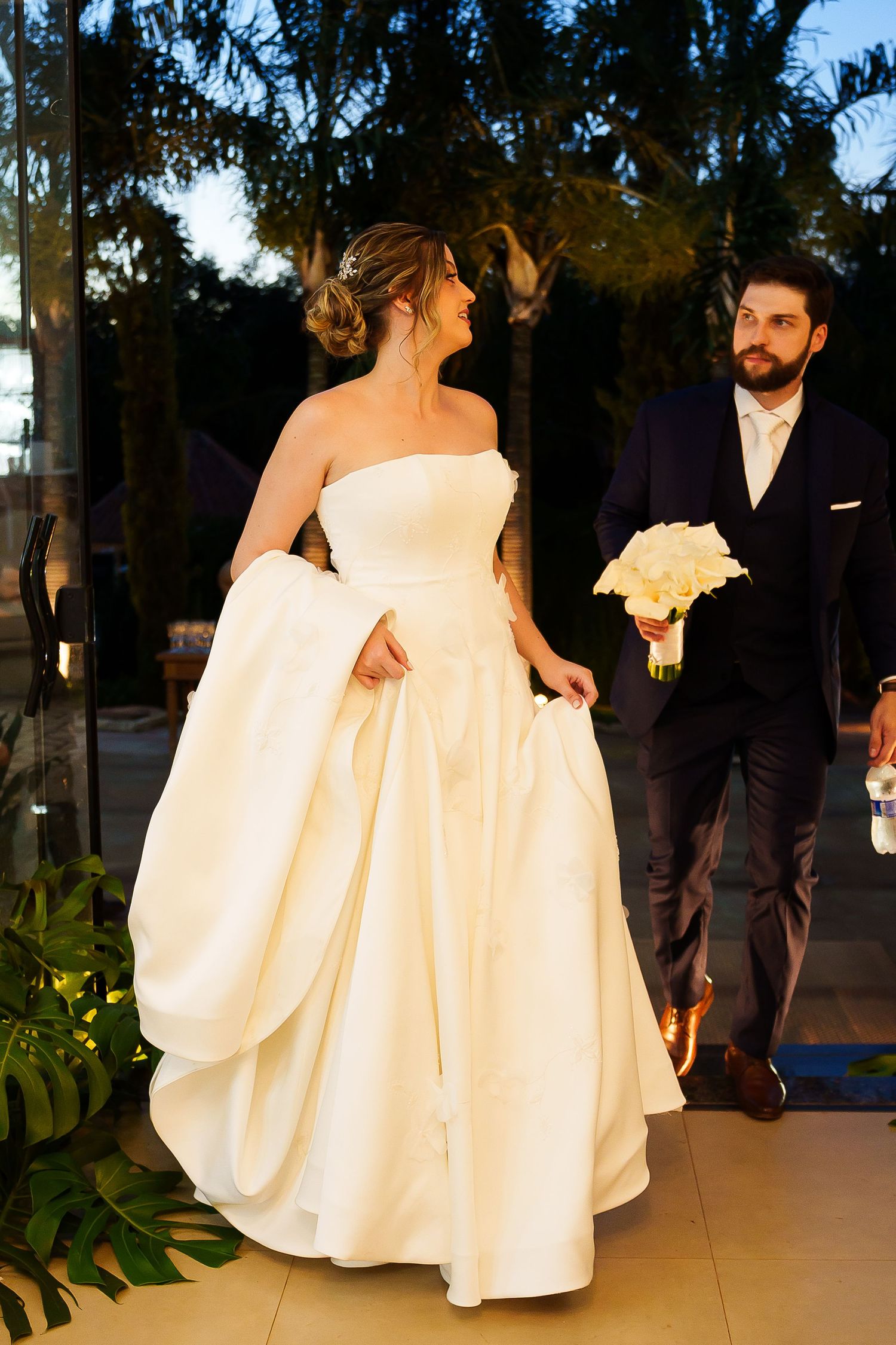 Elegant bride in strapless white ball gown walks with bearded groom in navy suit holding white bouquet.