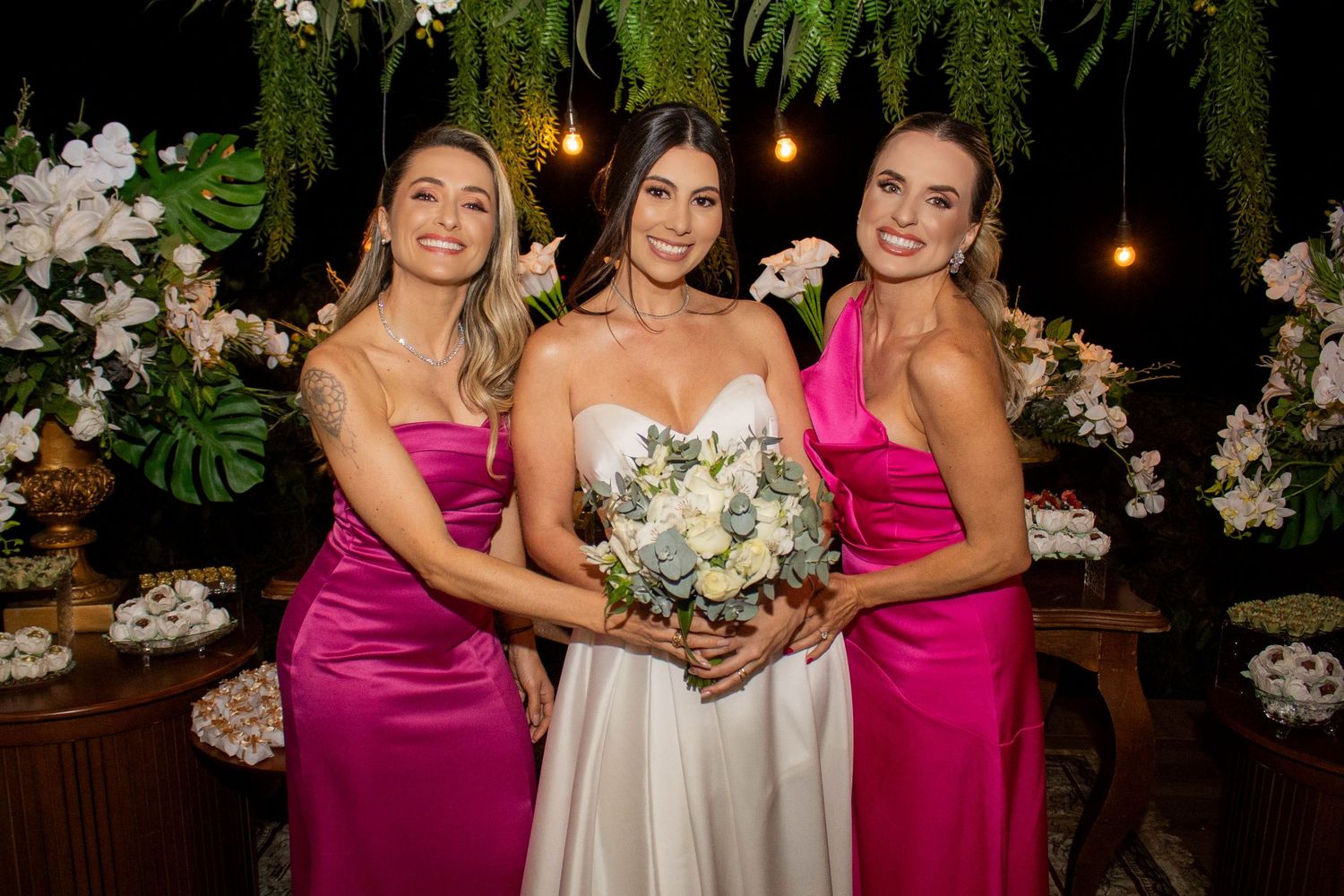 Bride in white gown holds bouquet flanked by two bridesmaids in pink satin dresses at floral wedding backdrop.